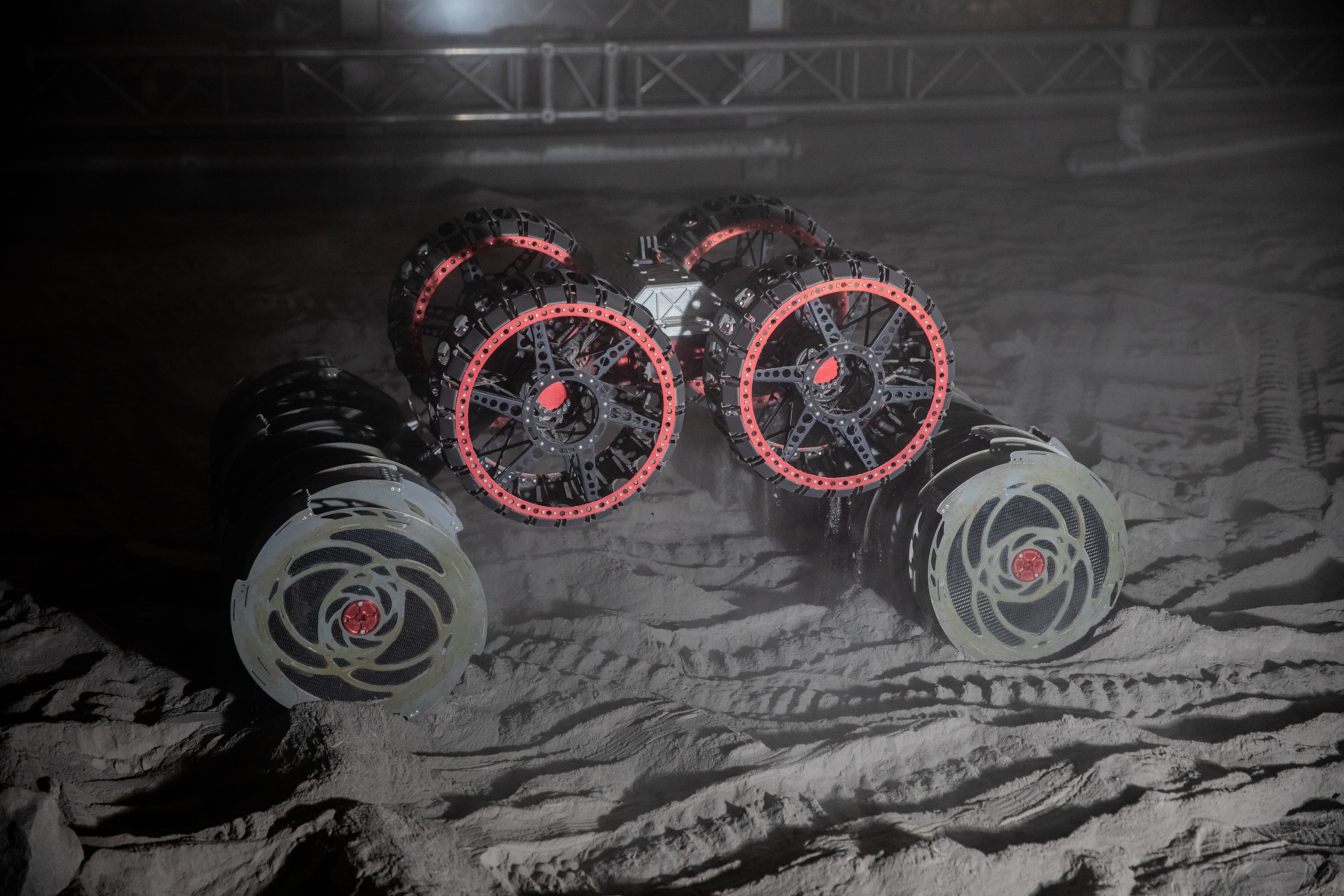 A wheeled robot navigates a sandy, dimly lit environment. Tracks in the sand indicate recent movement.