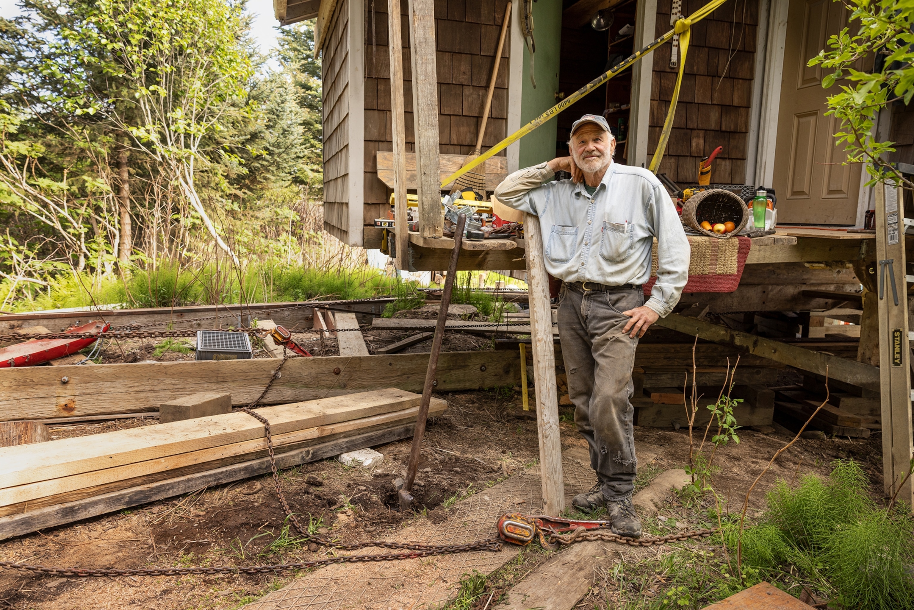 A man poses for a portrait in front of his small wooden house, which is propped up on pieces of wood and surrounded by tools and building materials.