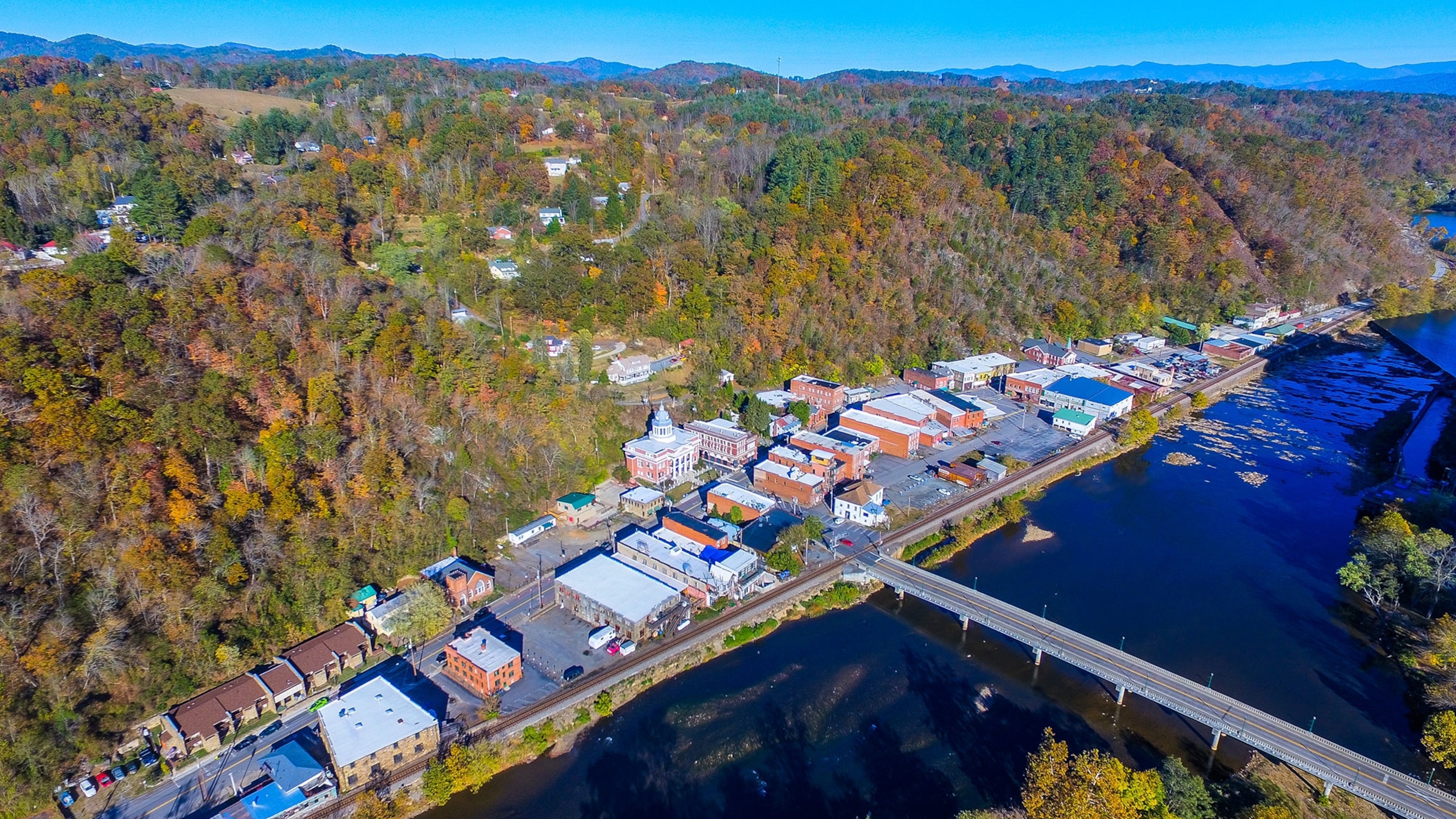 A an aerial view looks down on lush trees spread out into the distance in the background a small collection of buildings sits on the bank of a river with a bridge. Image Travel Changes Pending
