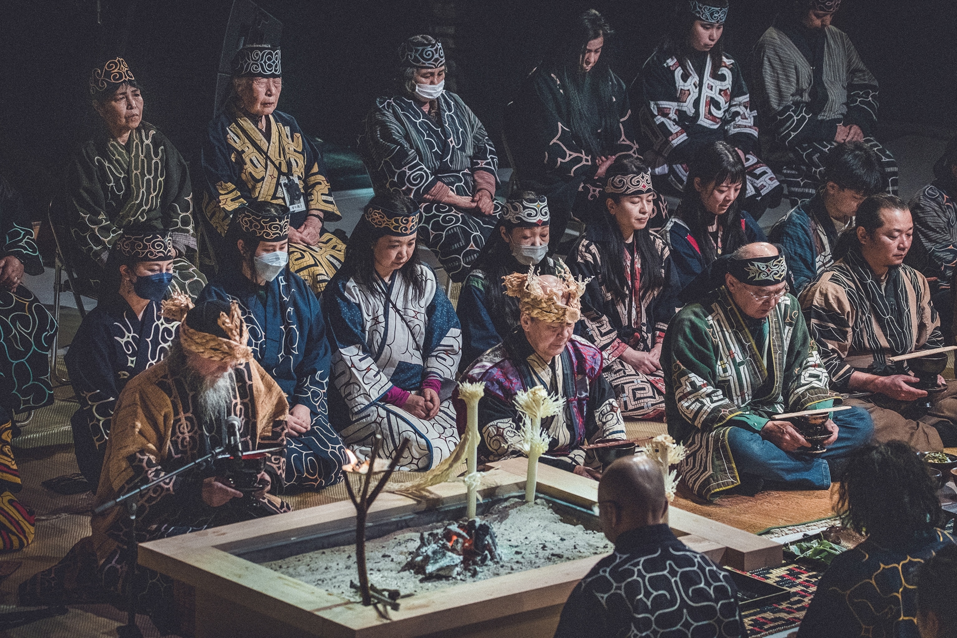The moody and dark interiors of a ceremonial room with rows of indigenous Ainu people sitting cross-legged on the floor in traditional clothing.