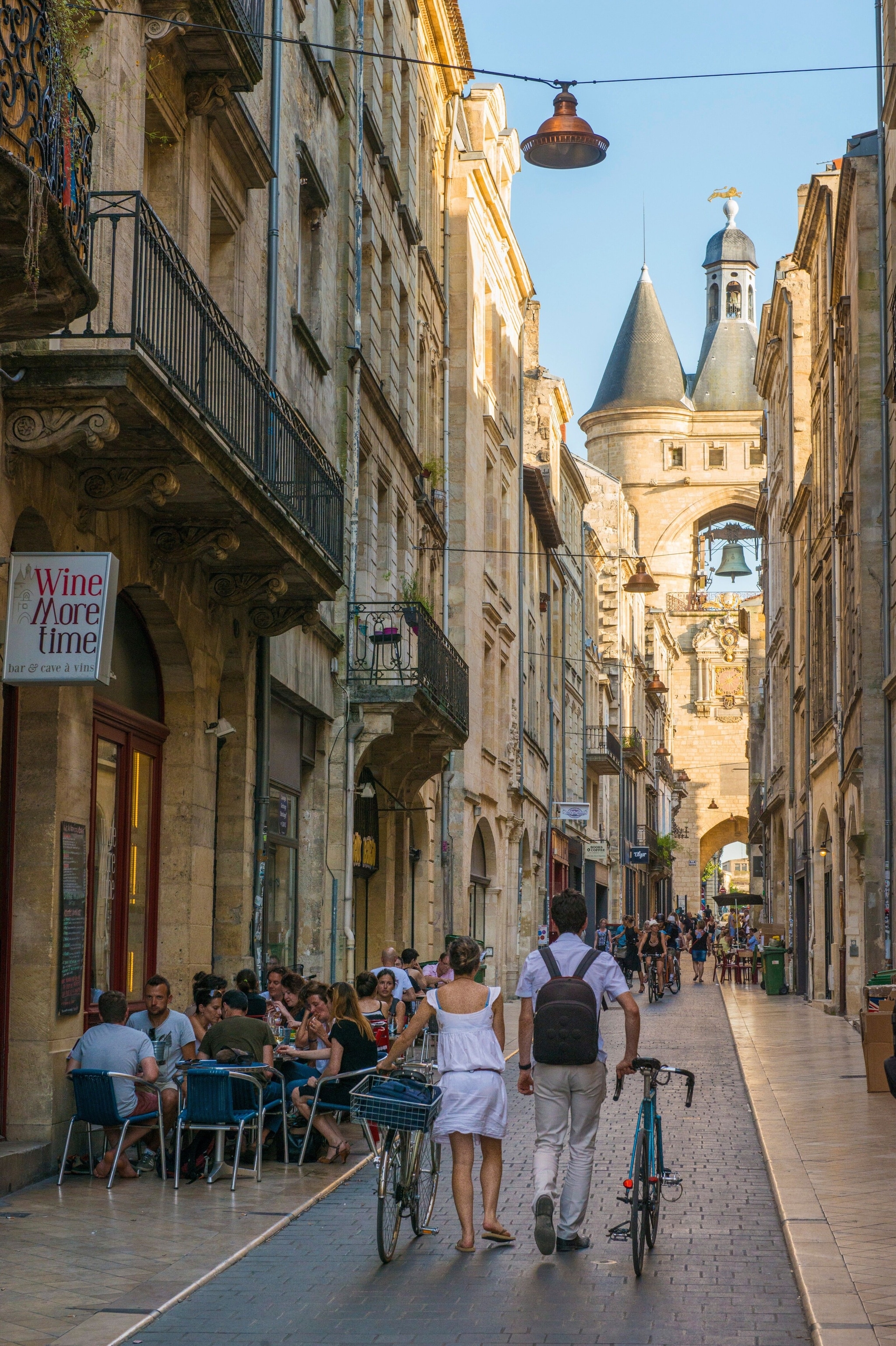 Rue Saint-James in Bordeaux. A man and a woman walk their bikes down the street. People sit at tables outside a cafe bar called 'Wine More time'. The buildings are tall and made of stone. A large bell tower is visible ahead.