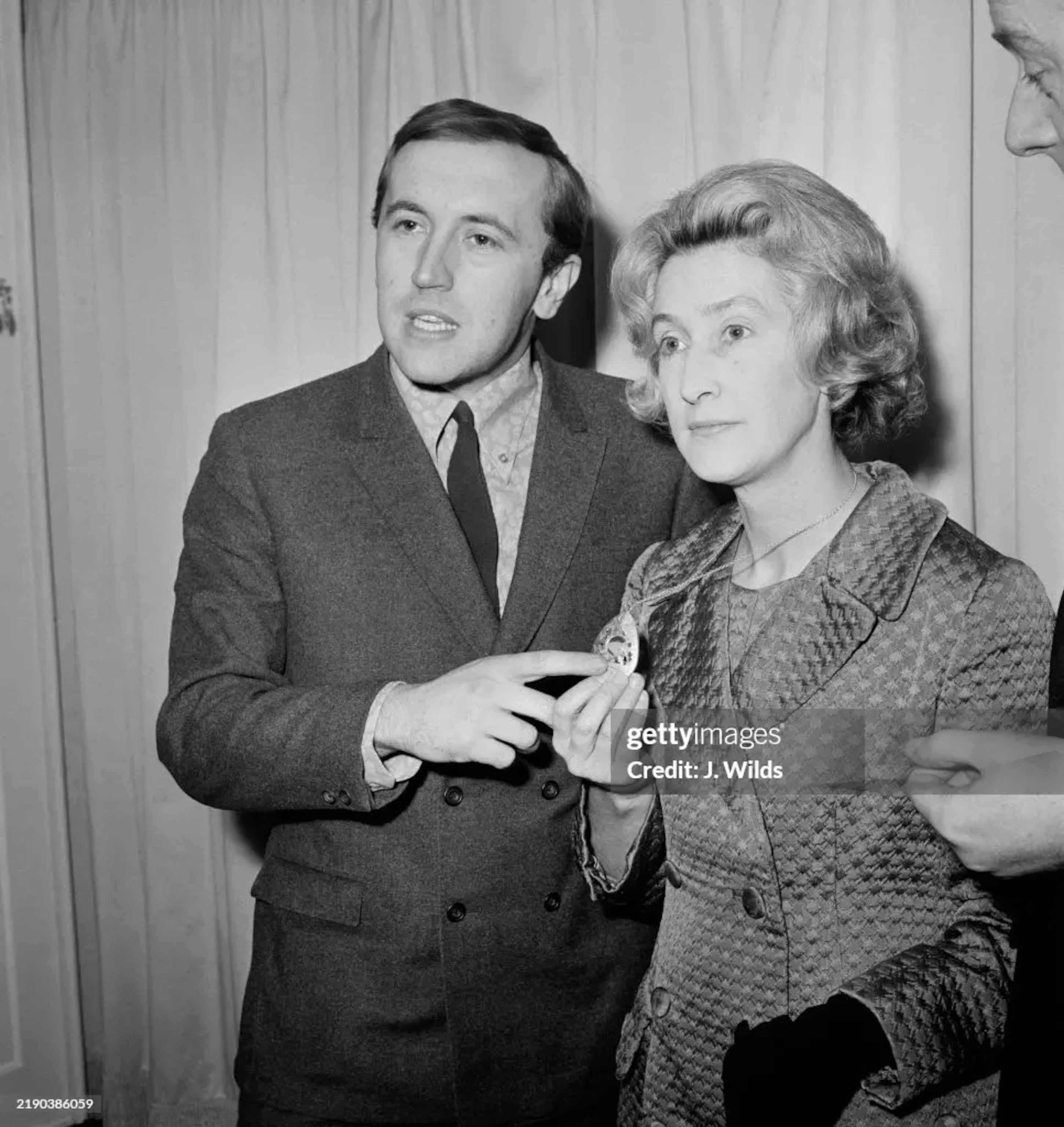 A man stands next to a woman and points to the necklace that she is wearing.