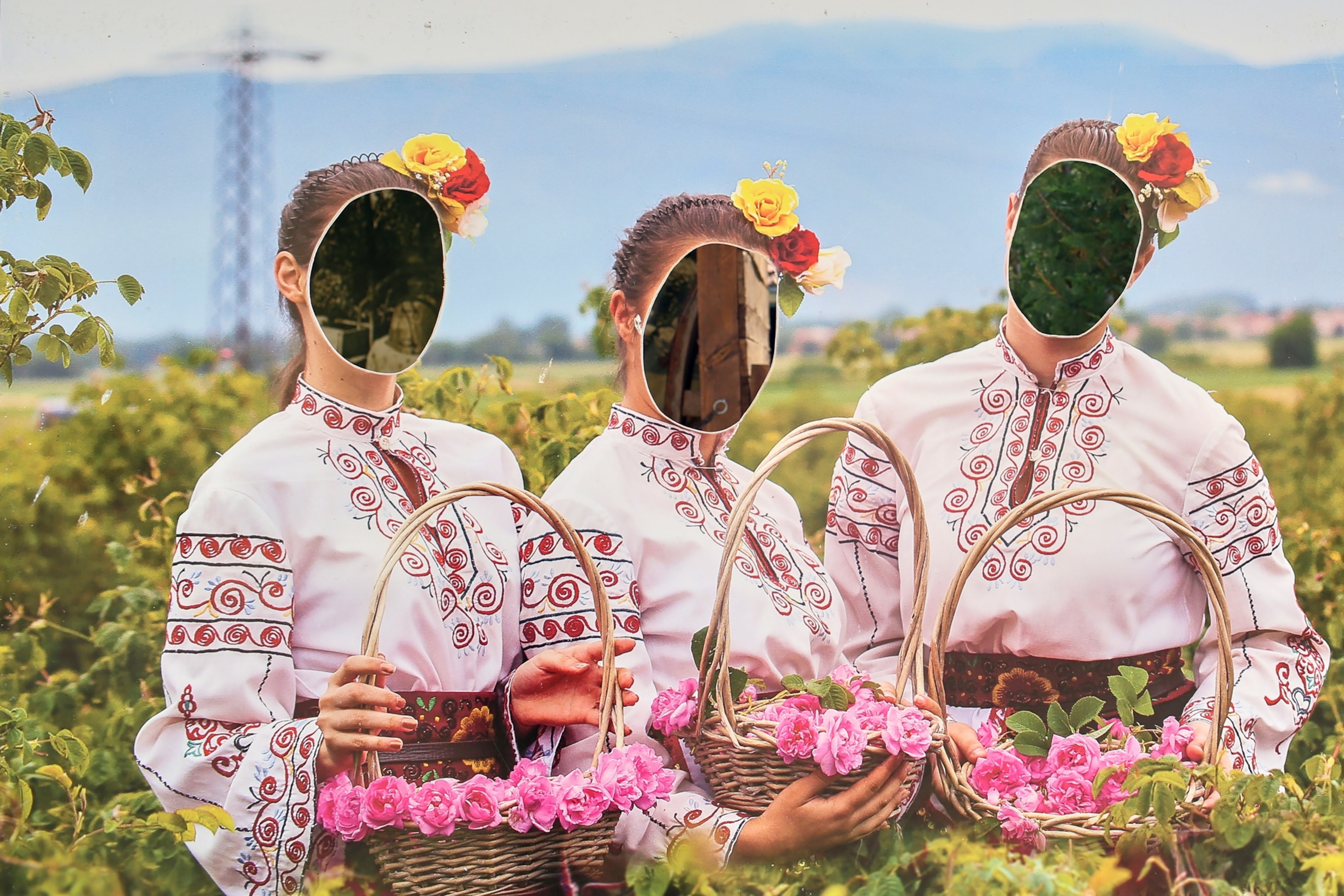 A cardboard cutout of three women in white embroidered dresses holding baskets of flowers. The faces are cut out of the cardboard.