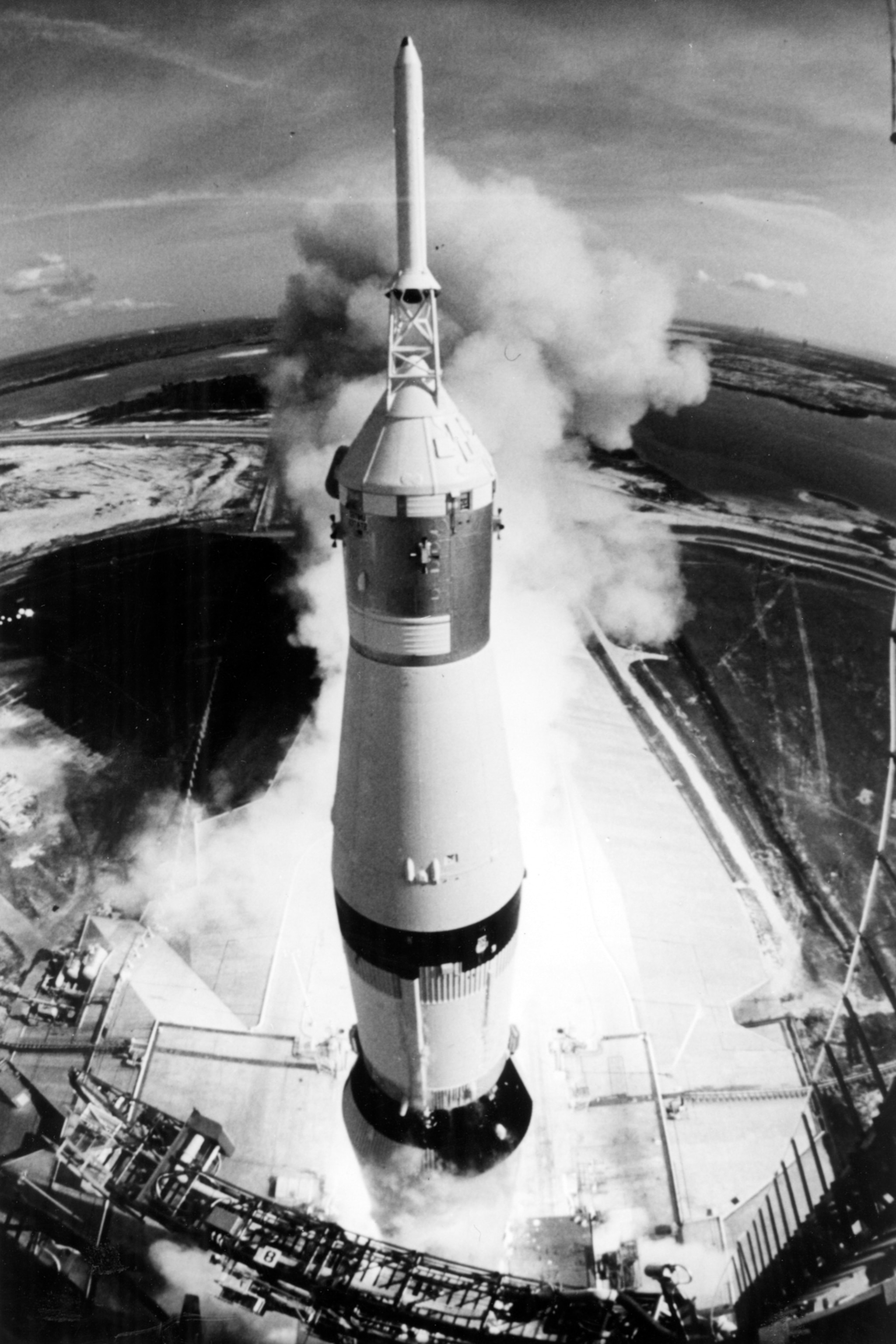 A large rocket launches vertically, emitting thick white smoke. The ground below shows a launch pad and surrounding landscape