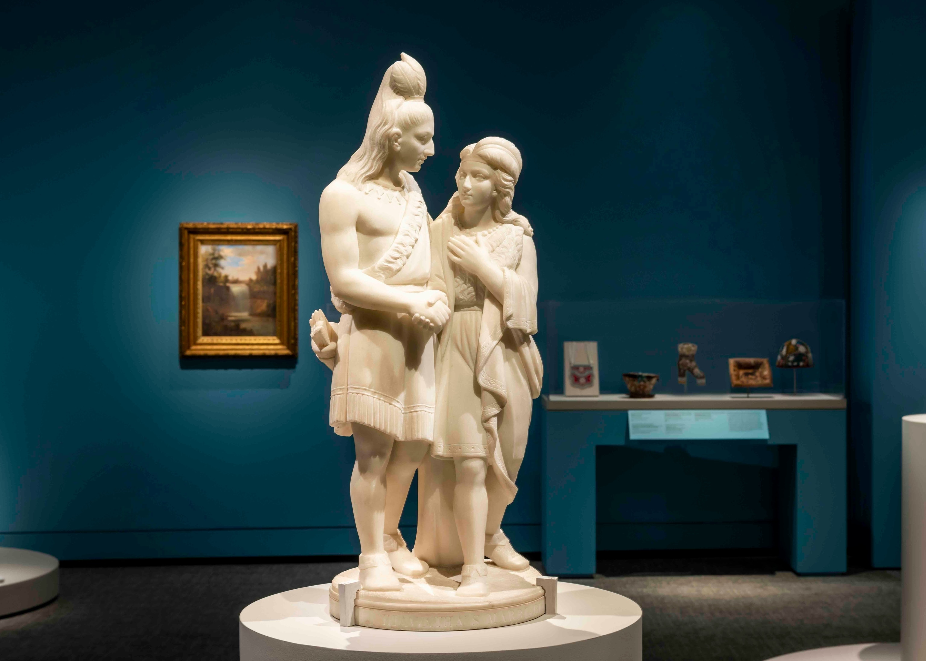 A white marble statue of two embracing figures stands in the center of a room with other artifacts and works of art around