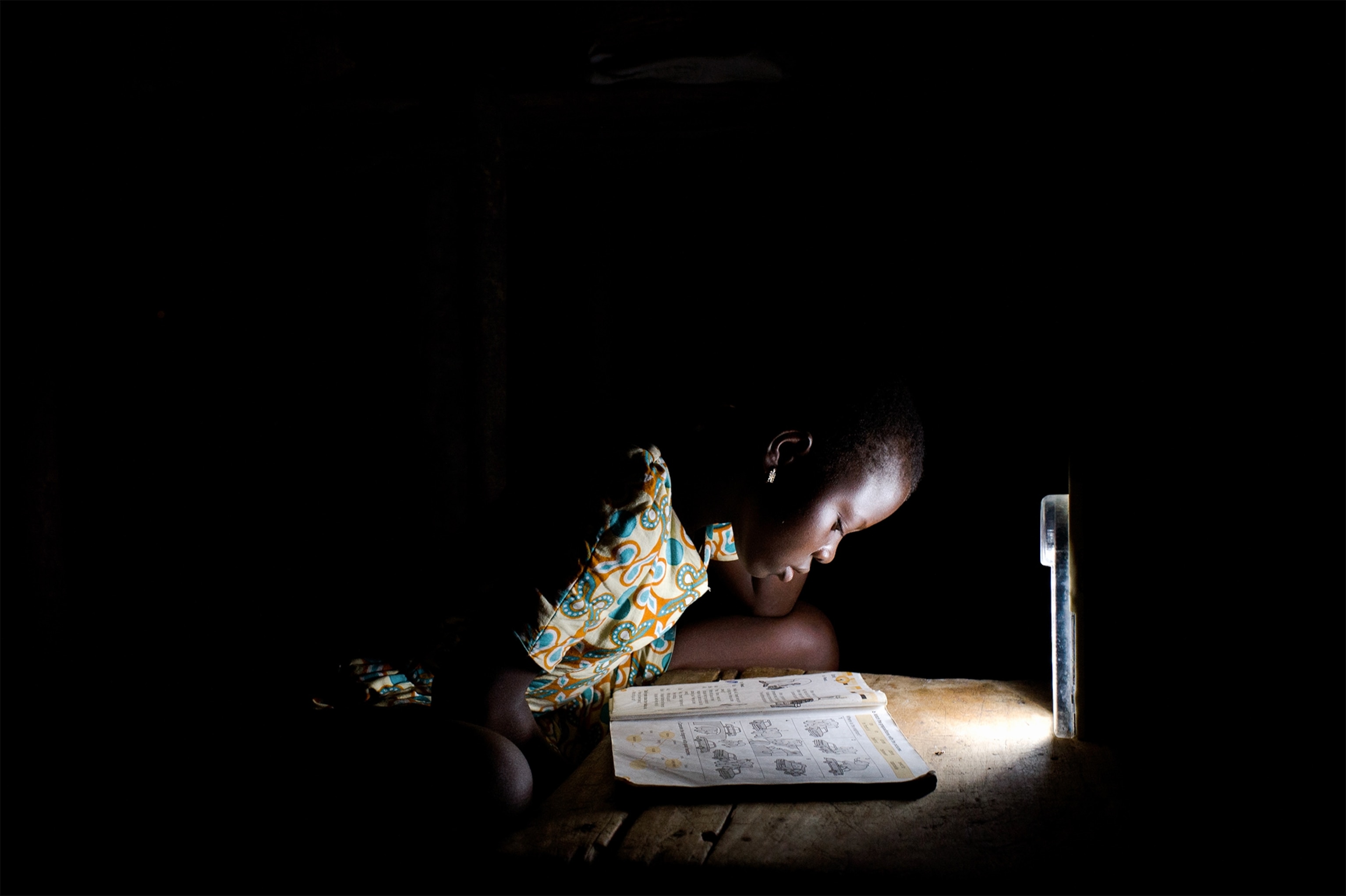 a Ghanian girl reading an english textbook by a small light in the dark.
