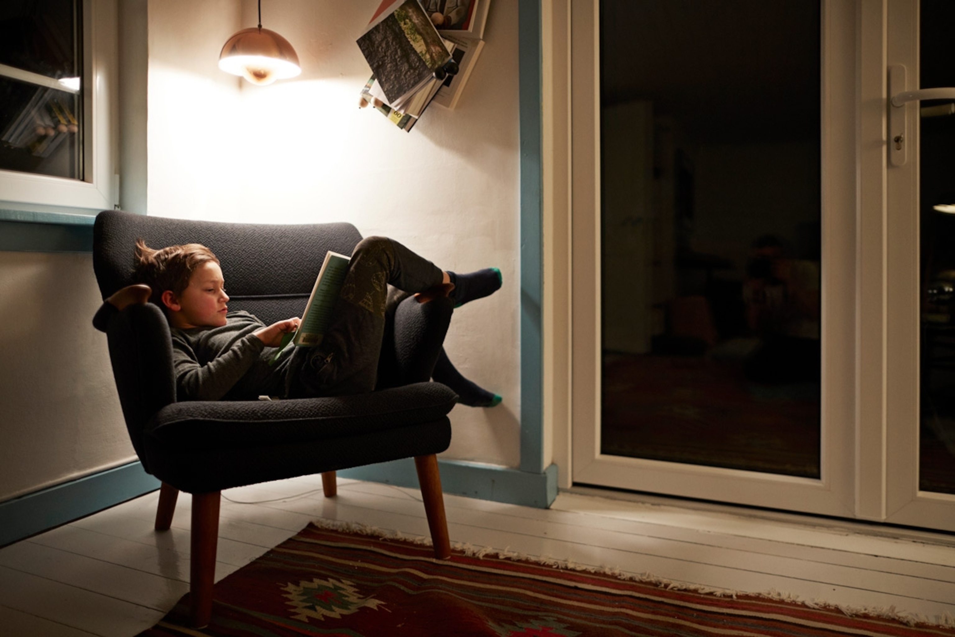 Boy reading book under lamp in armchair at night. Copenhagen, Denmark.