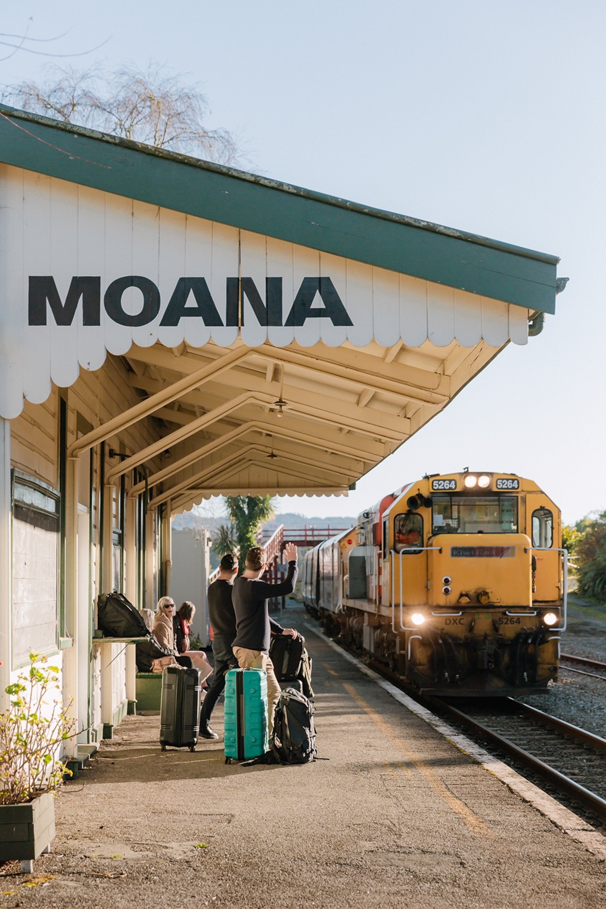 A wooden train station with a scalloped roof and passengers with suits waving in the TranzAlpine train.