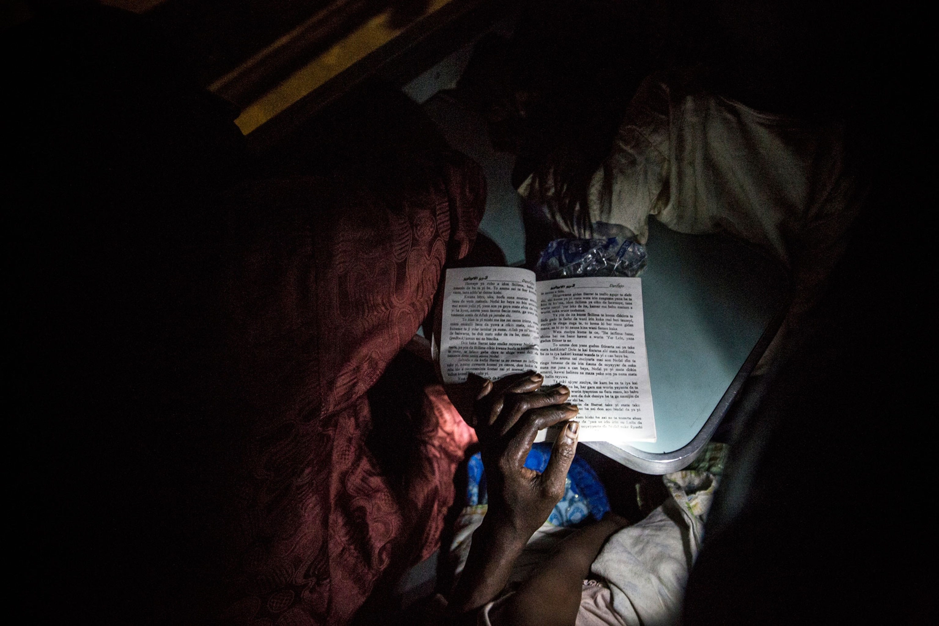 On a train from Lagos to Kano, a Hausa woman reads a romance novel with the flashlight of her phone.