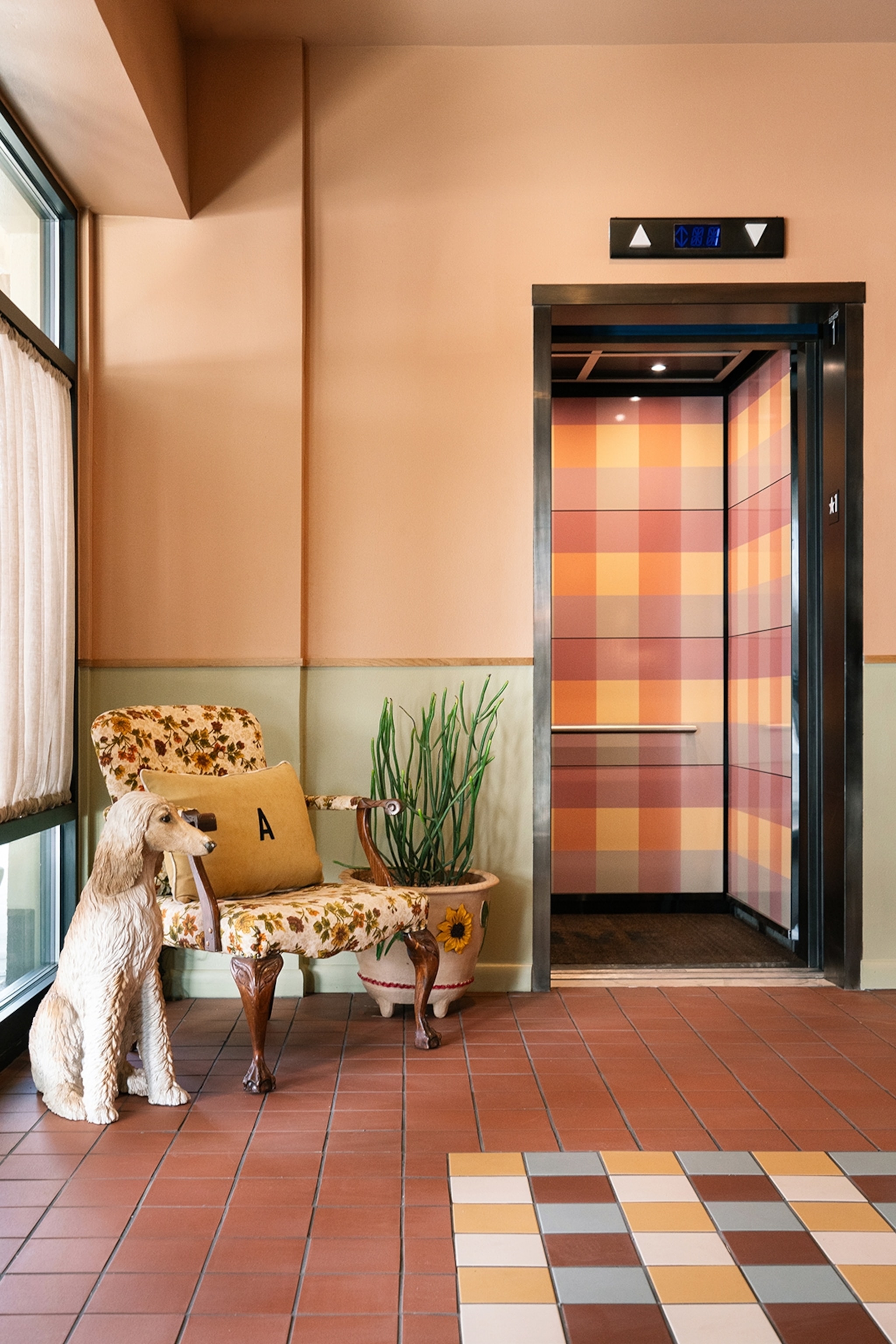 A retro-tiled hallway in a hotel with a patterned elevator and an armchair by the entrance.