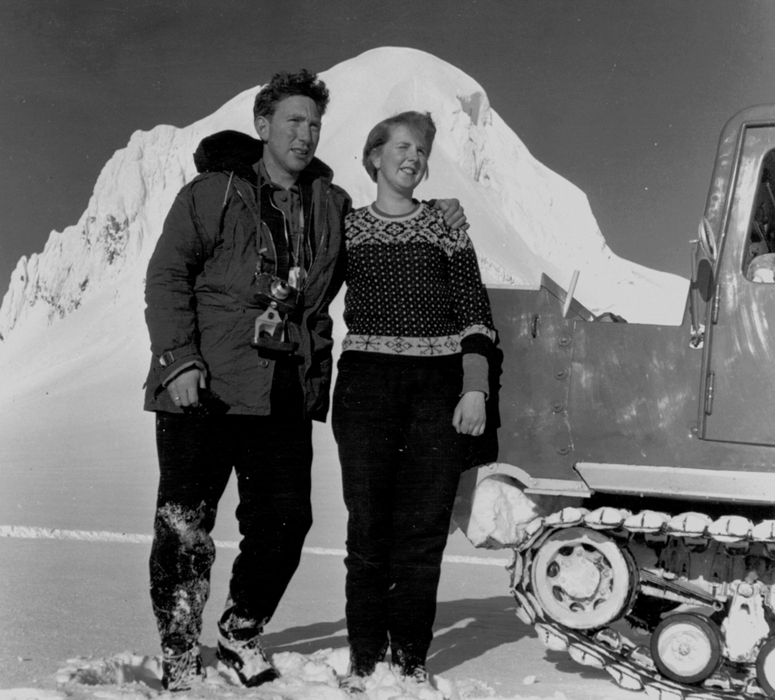 A man wearing a winter parka with a camera around his neck stands with a woman wearing a sweater, behind them stands a snowy mountain peak.