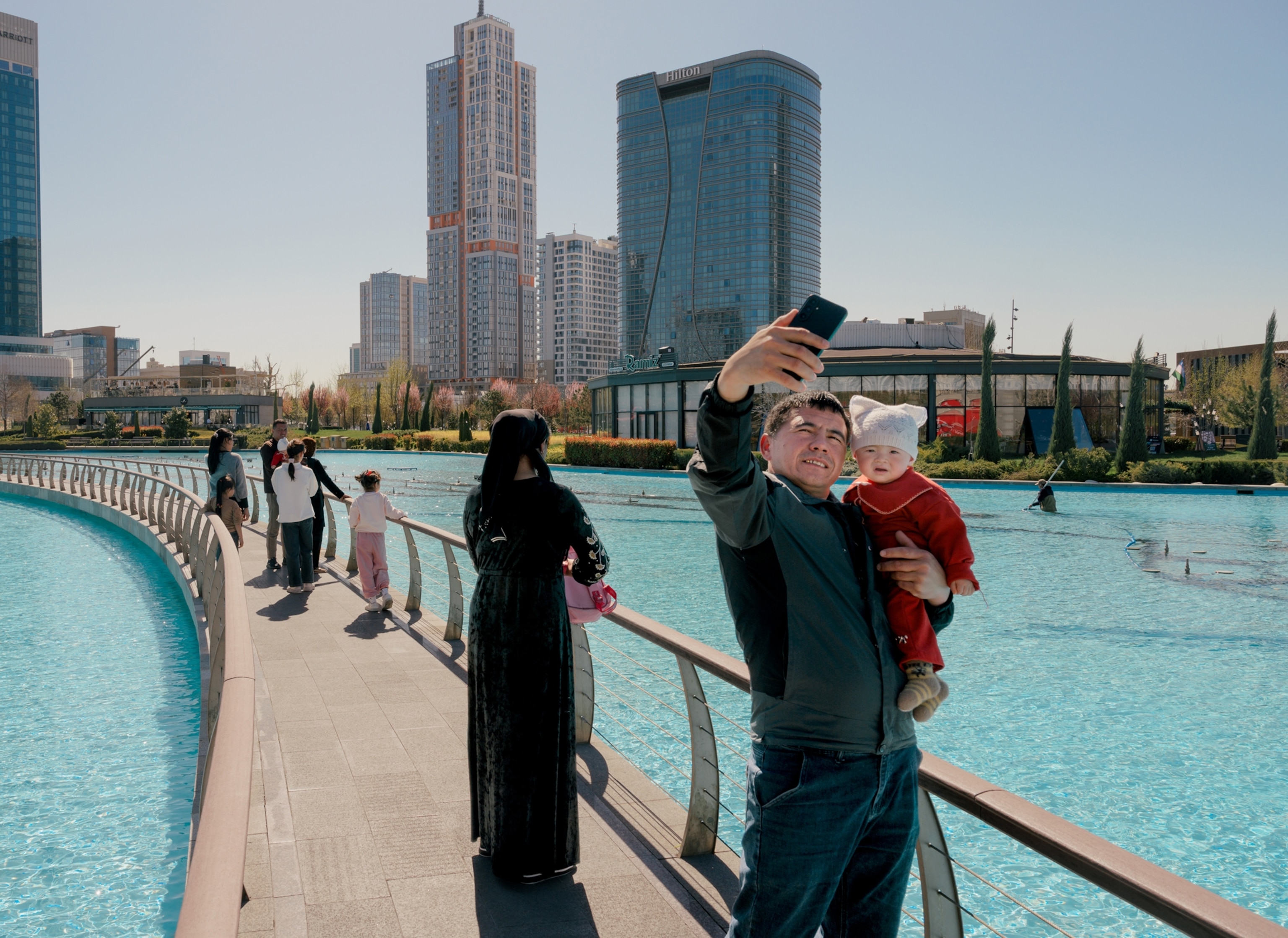 A sidewalk stretches across a man-made body of water with tall buildings in the background. Several people walk across the bridge and a man in the foreground takes a selfie while holding his infant daughter.