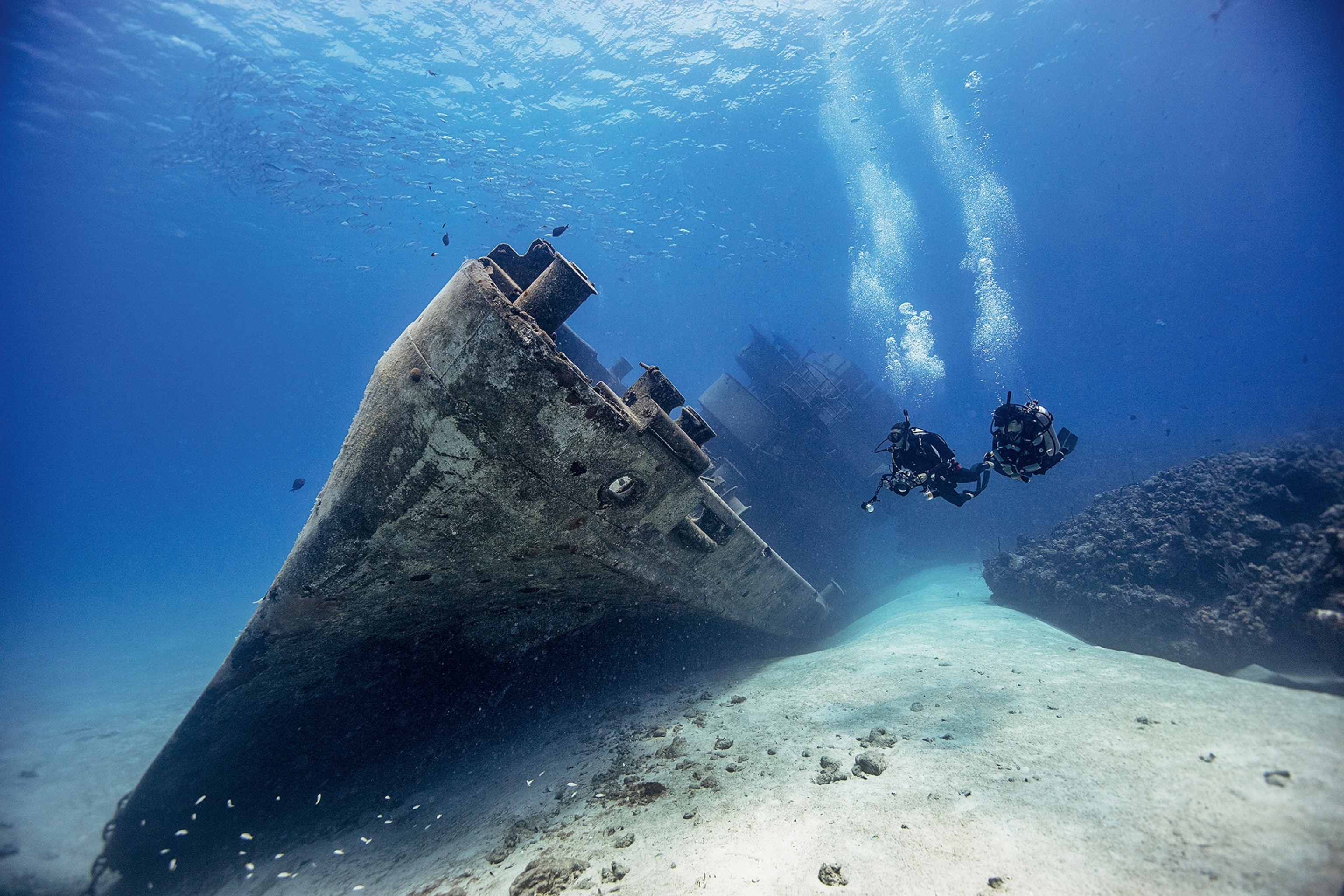 A wide underwater shot of a massive shipwreck with a swarm of small fish above and two divers looking at the ship's side.