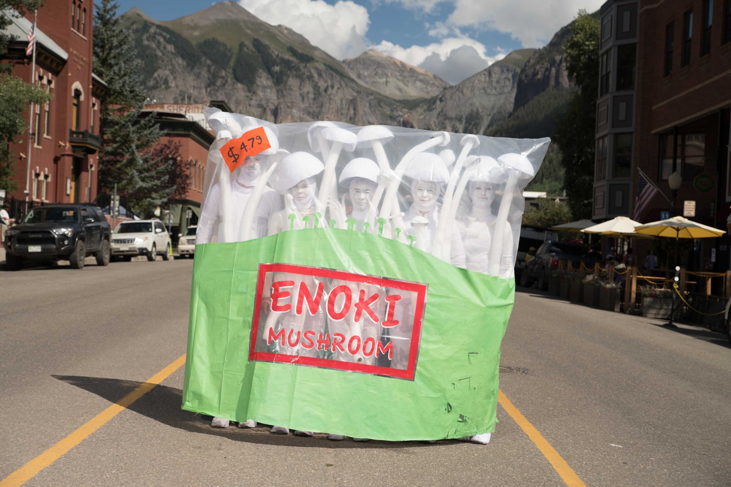A group of people huddle together in cosumes with white face paint body suits and mushroom cap hats while holding human size long mushroom cap props.
