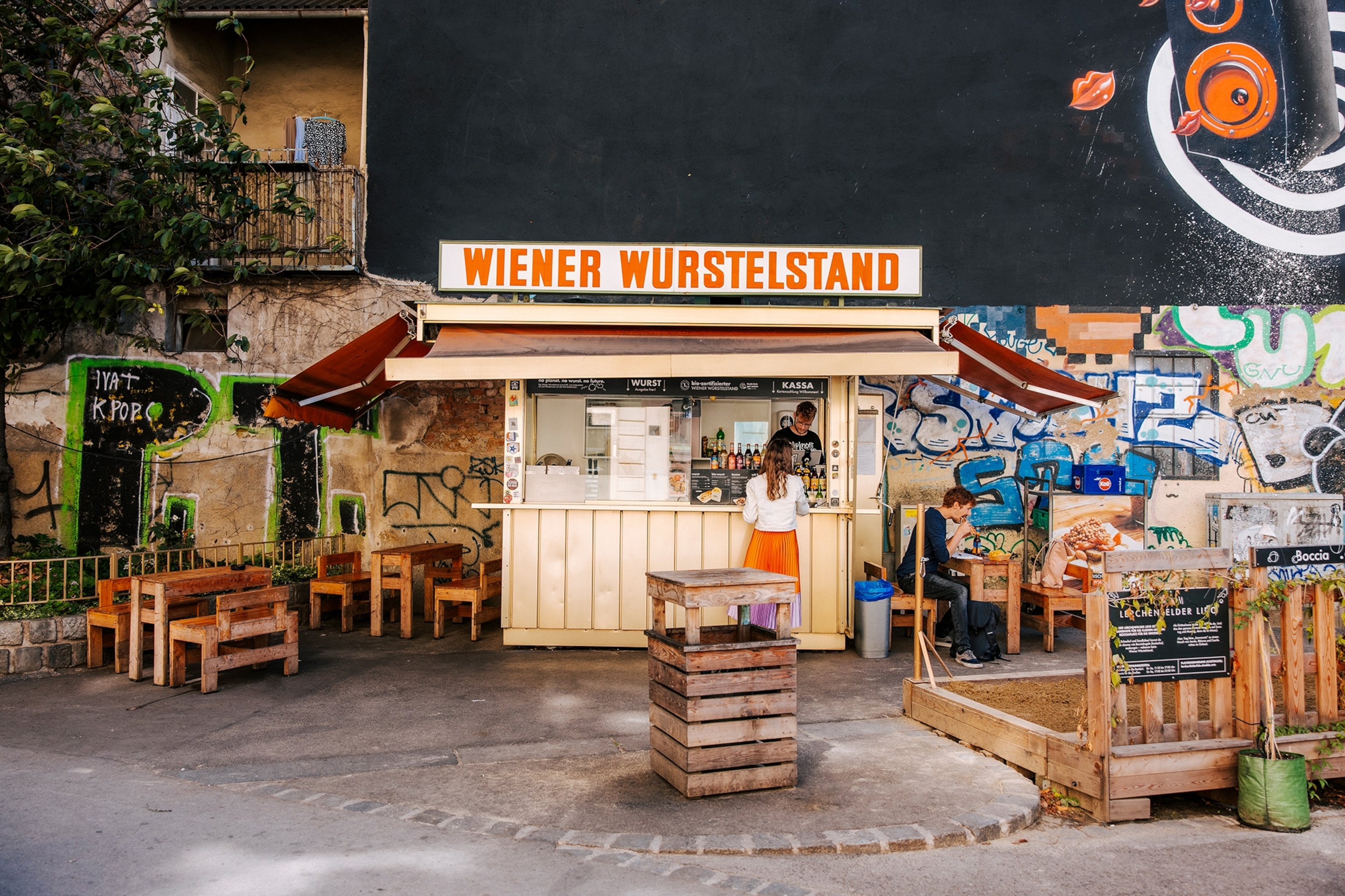 A courtyard kiosk selling sausages surrounded by simple wooden chairs.