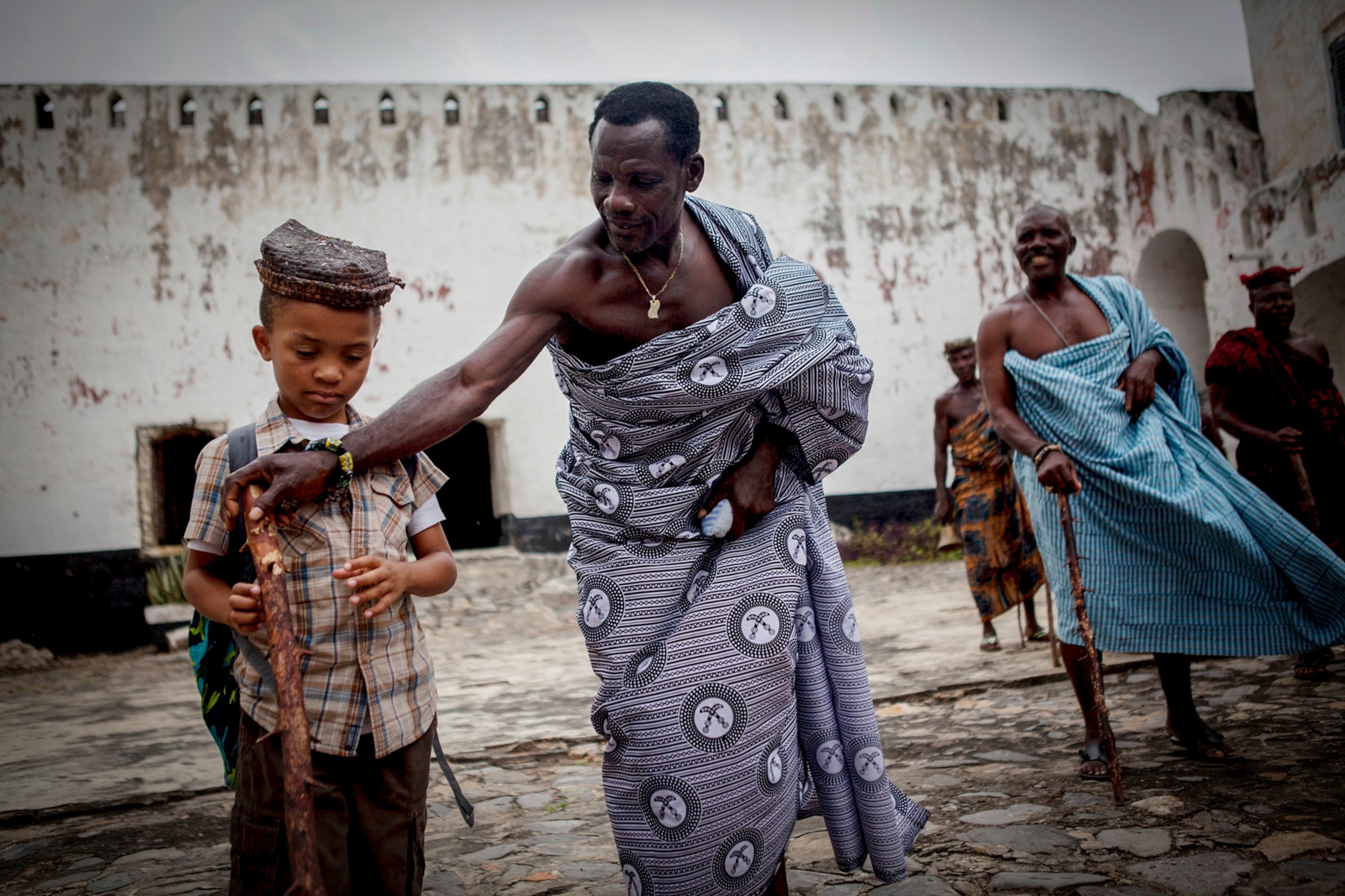 an elder adjusts a young tourist’s grip on a traditional staff in Anomabo, Ghana