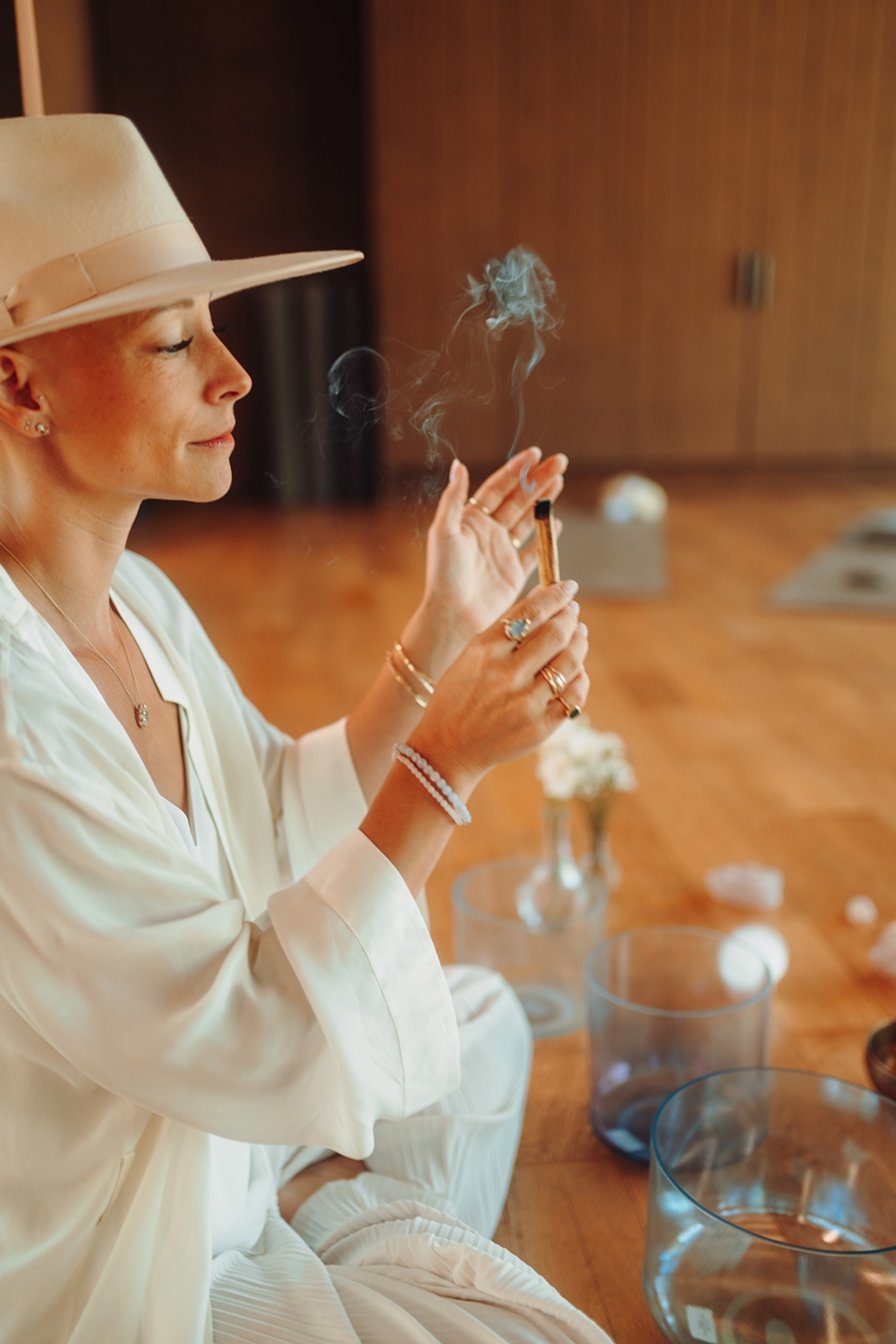 A woman with a fedora sitting on the wooden floor of a wellness studio with three bowls of varying size in front of her as she burns incense with her eyes closed.
