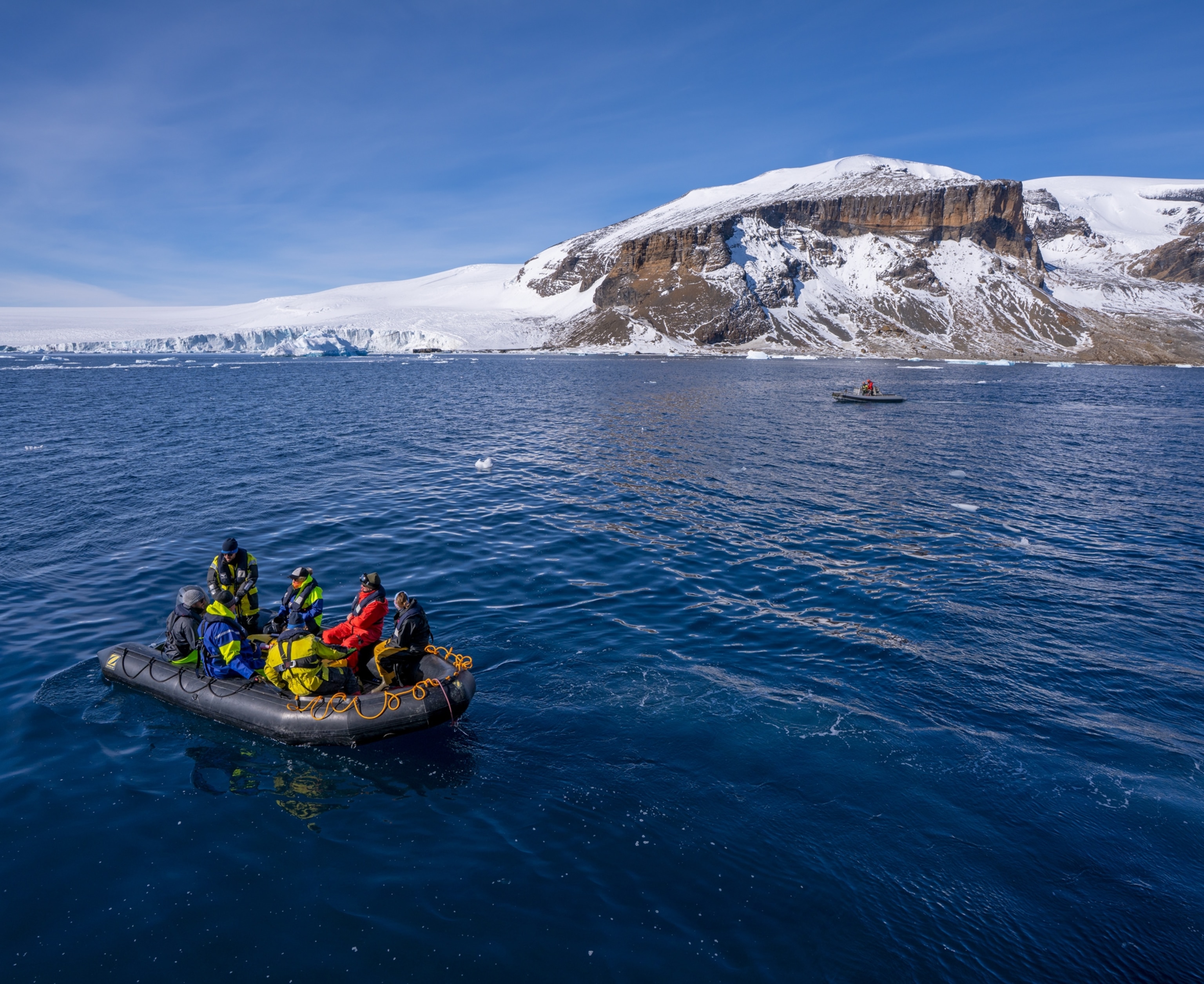 Group of people in colorful coats on the inflatable boat at very blue water with icy rocks on the background.