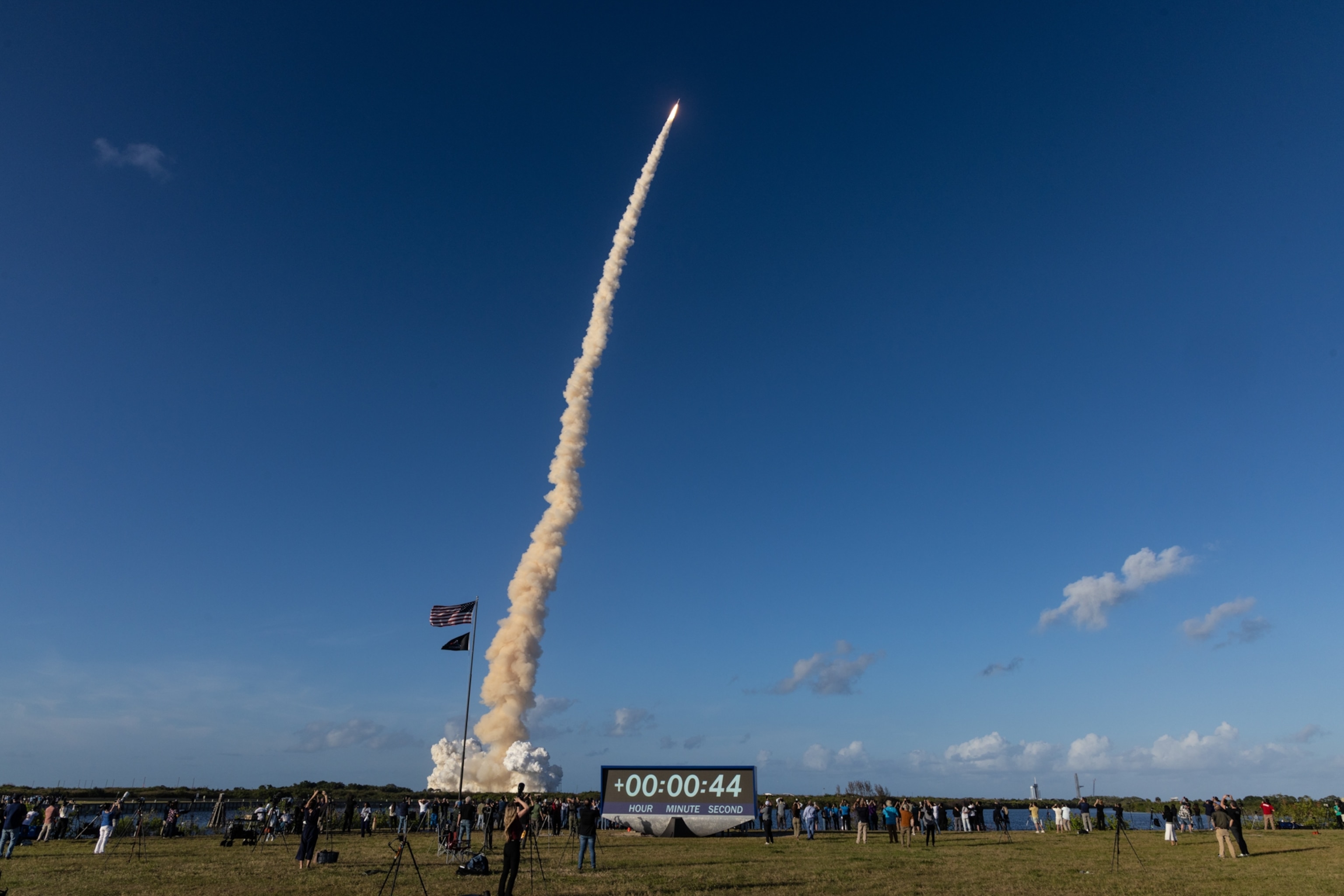 A rocket ascends into a clear blue sky, leaving a white smoke trail. A crowd watches from below, with flags and a countdown clock reading 00:00:44.