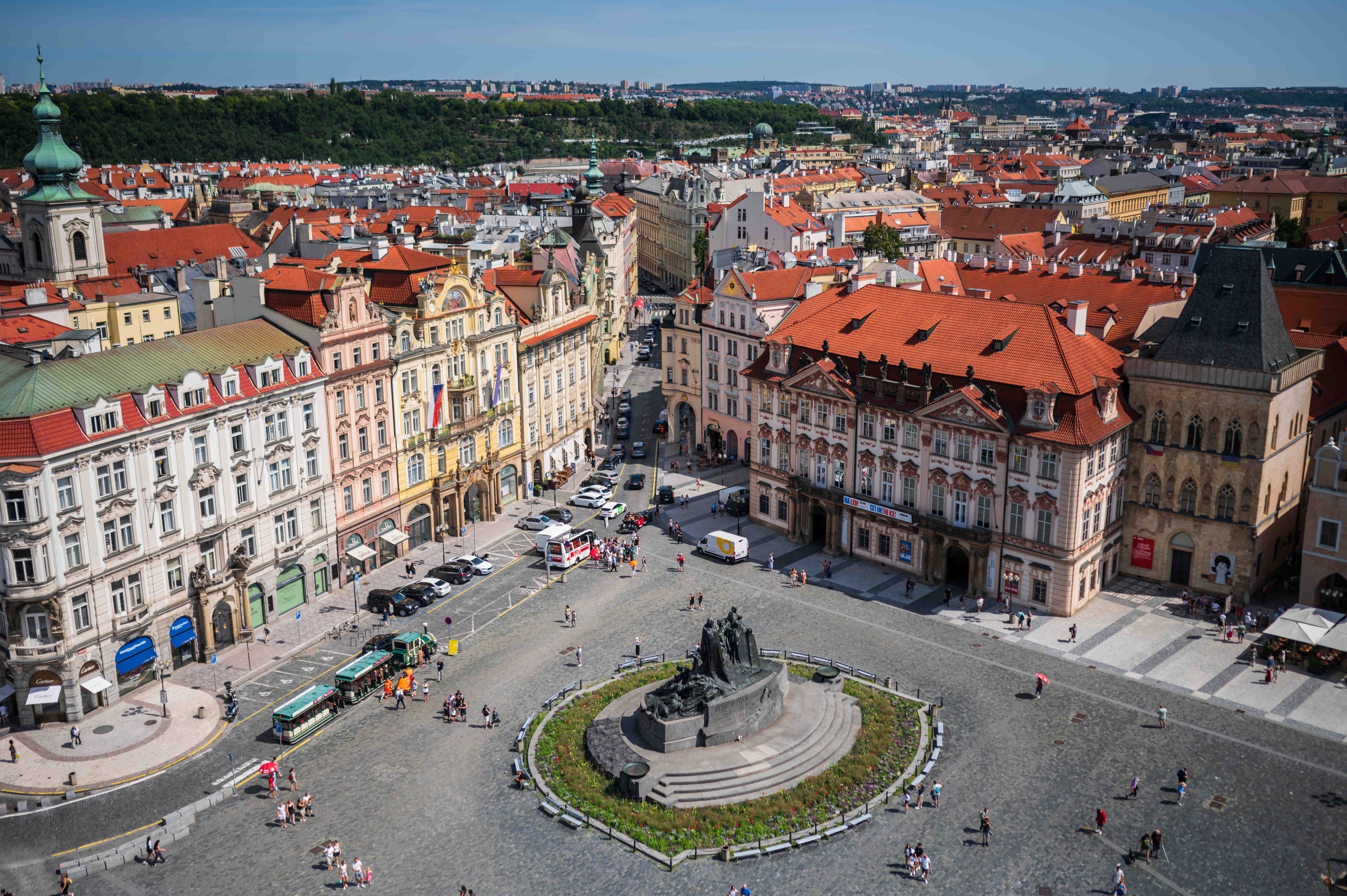 erial view of Old Town Square in Prague, Czech Republic, showing colorful historic buildings with red-tiled roofs, the Jan Hus monument in the center, and people walking through the cobblestone plaza.