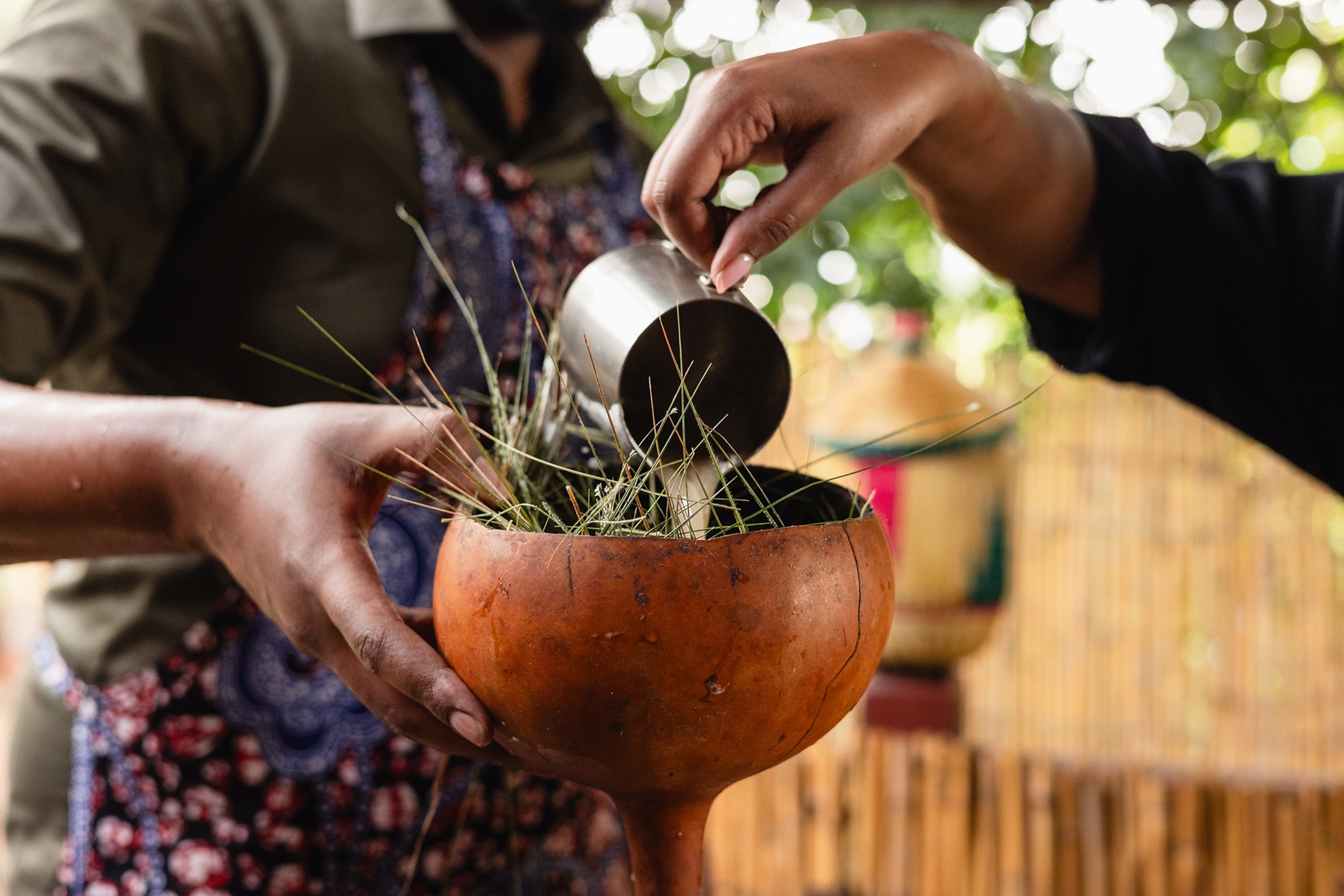 A focused shot of someone pouring banana beer from a milk steaming cup into a dried gourd vessel during a workshop.
