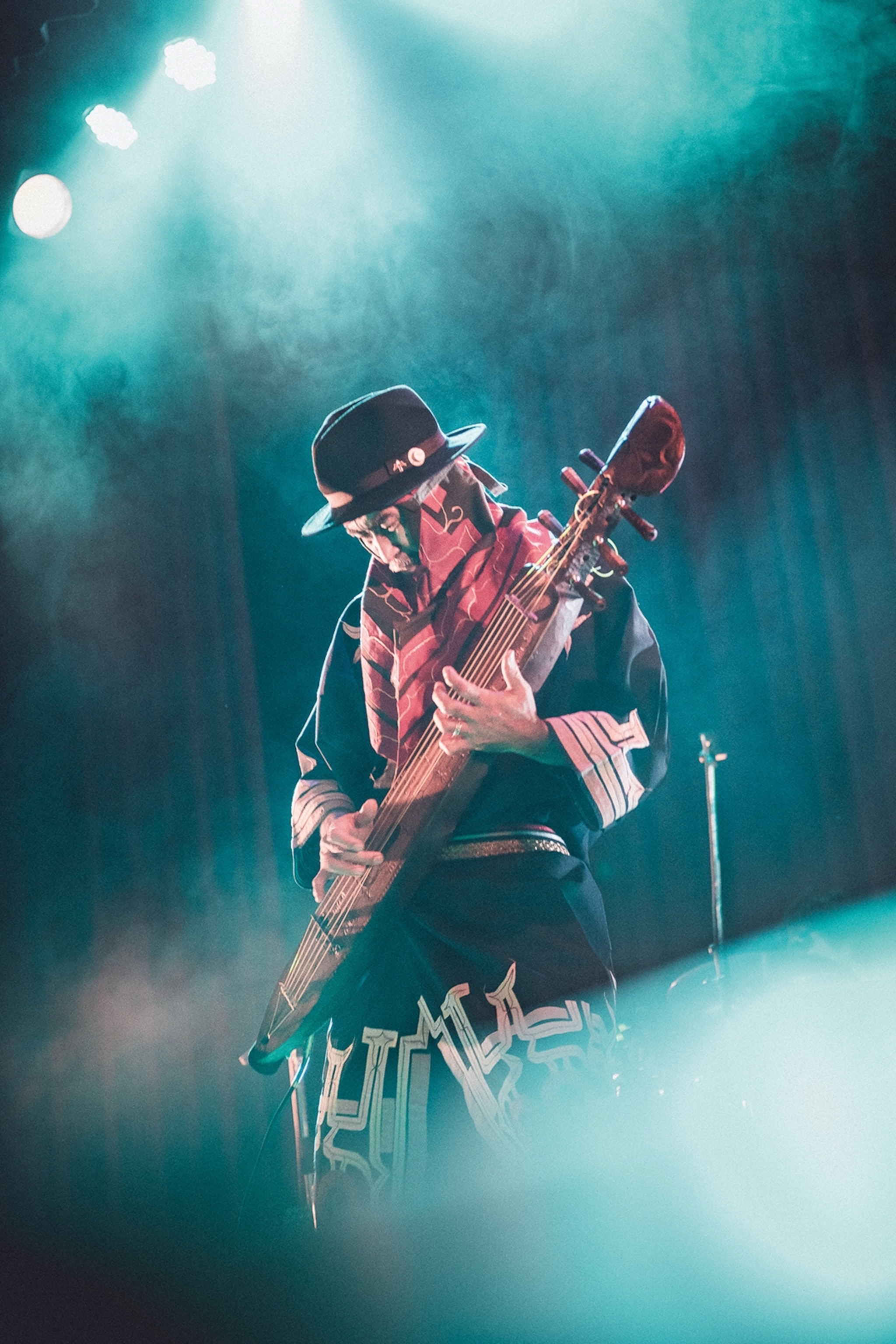 A dynamic concert shot of an eldery man in fedora on stage, playing a traditional, guitar-like harp.