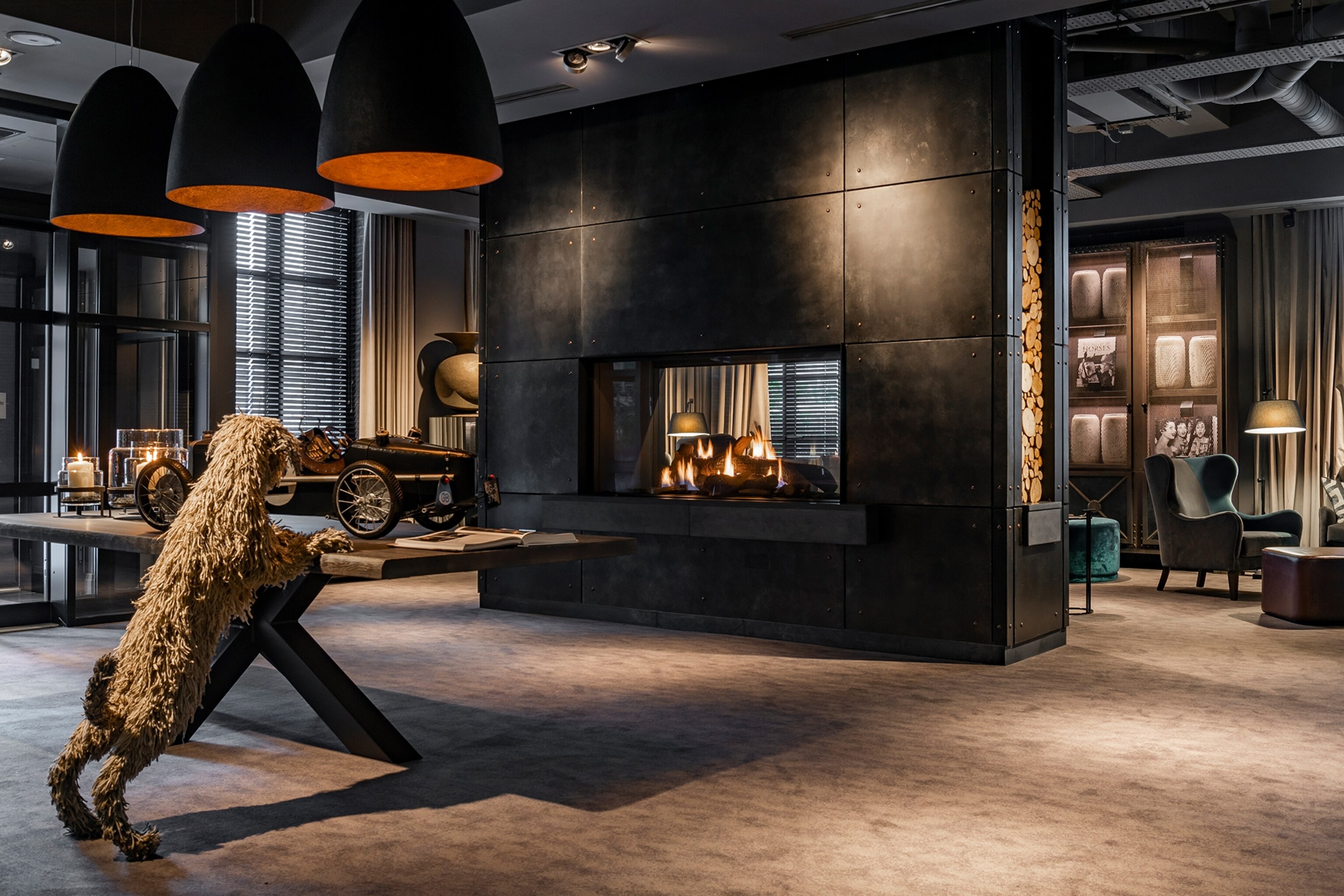 A moody and dark hotel lobby with a room-dividing fireplace and velvet arm chairs.