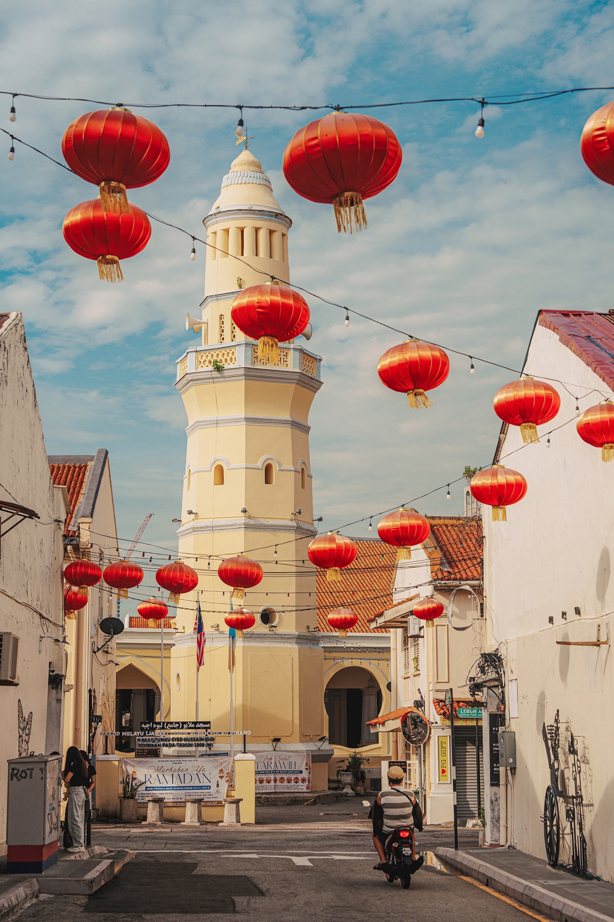 A narrow street lined with historic buildings is decorated with red lanterns hanging overhead, with a cream-colored tower rising in the center beneath a blue sky.