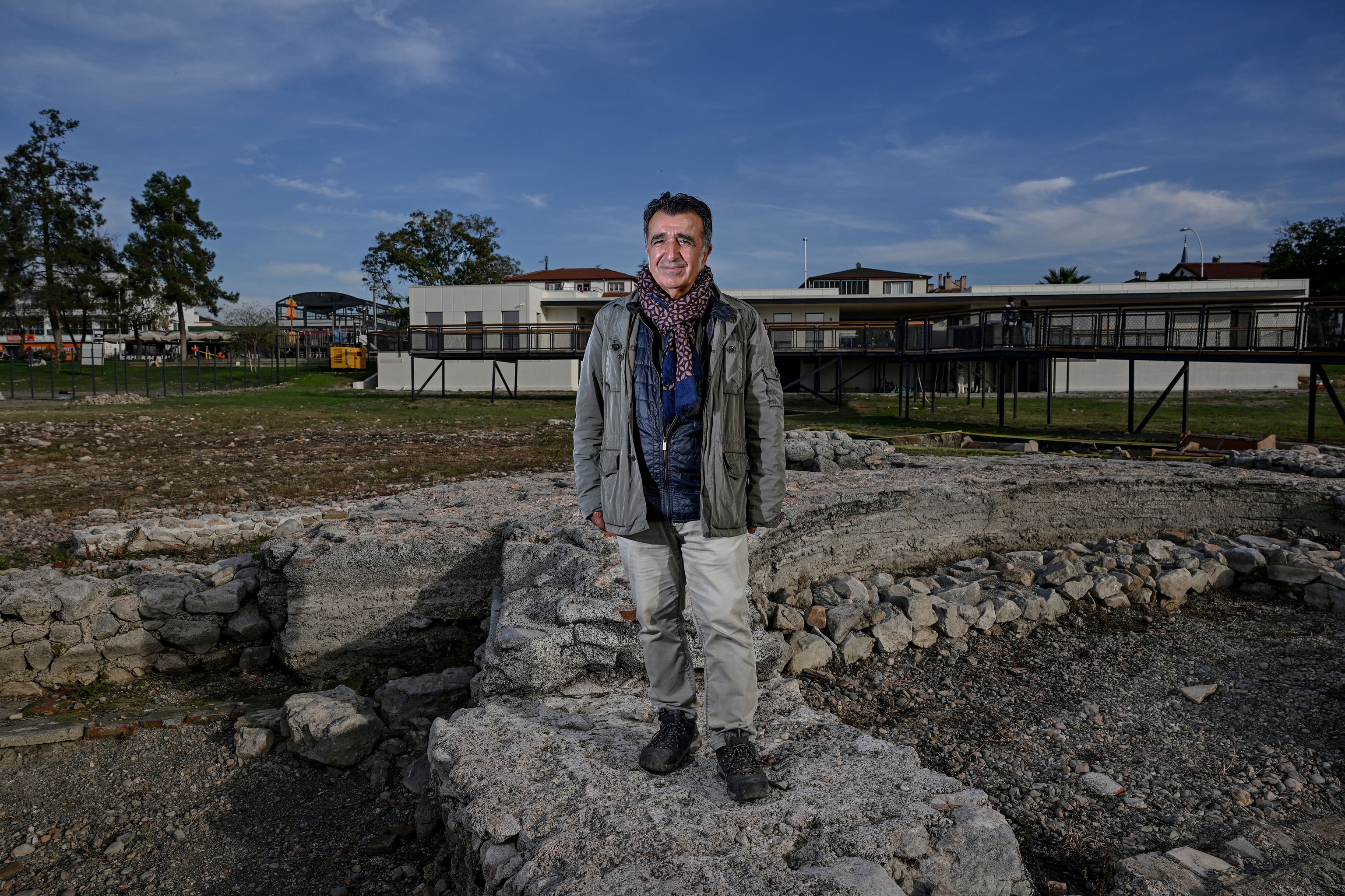 A man wearing a gray jacket and a sweater stands on a rock and smiles for a photo.