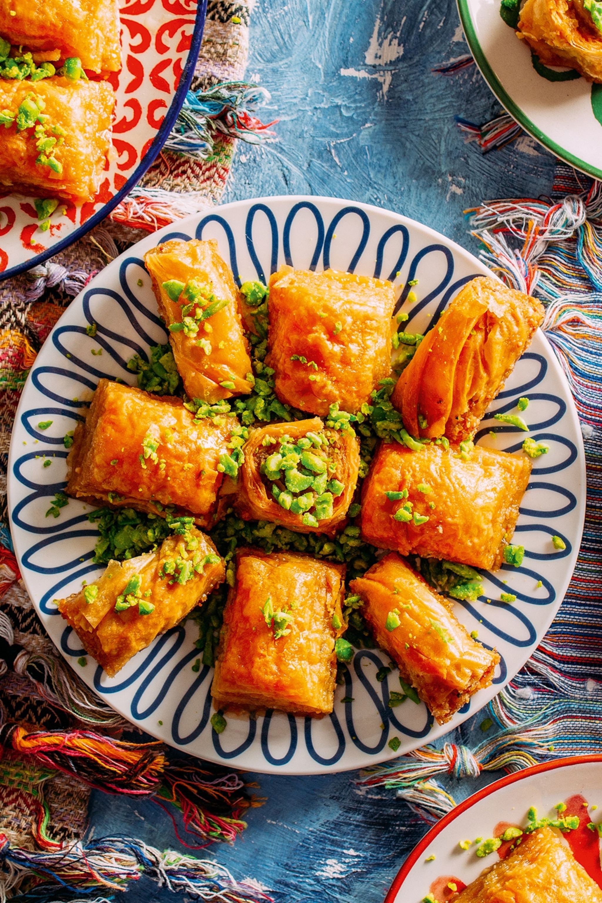 Plates of baklava pastry topped with vibrant pistachio on a traditionally weaved table cloth.