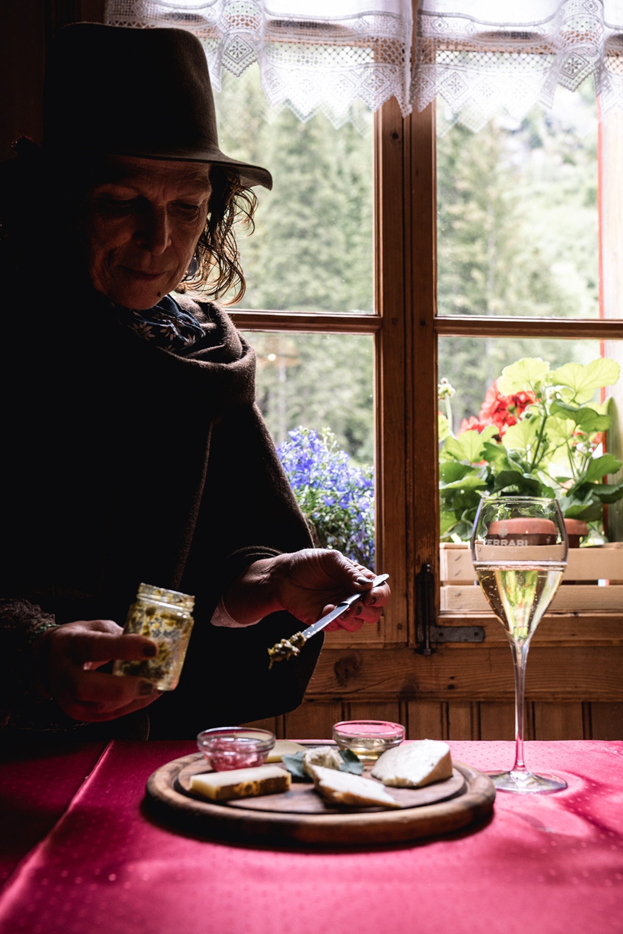 A woman in felt hat sitting by a rustic window at a dining table with bread, spreads and a glass of prosecco.