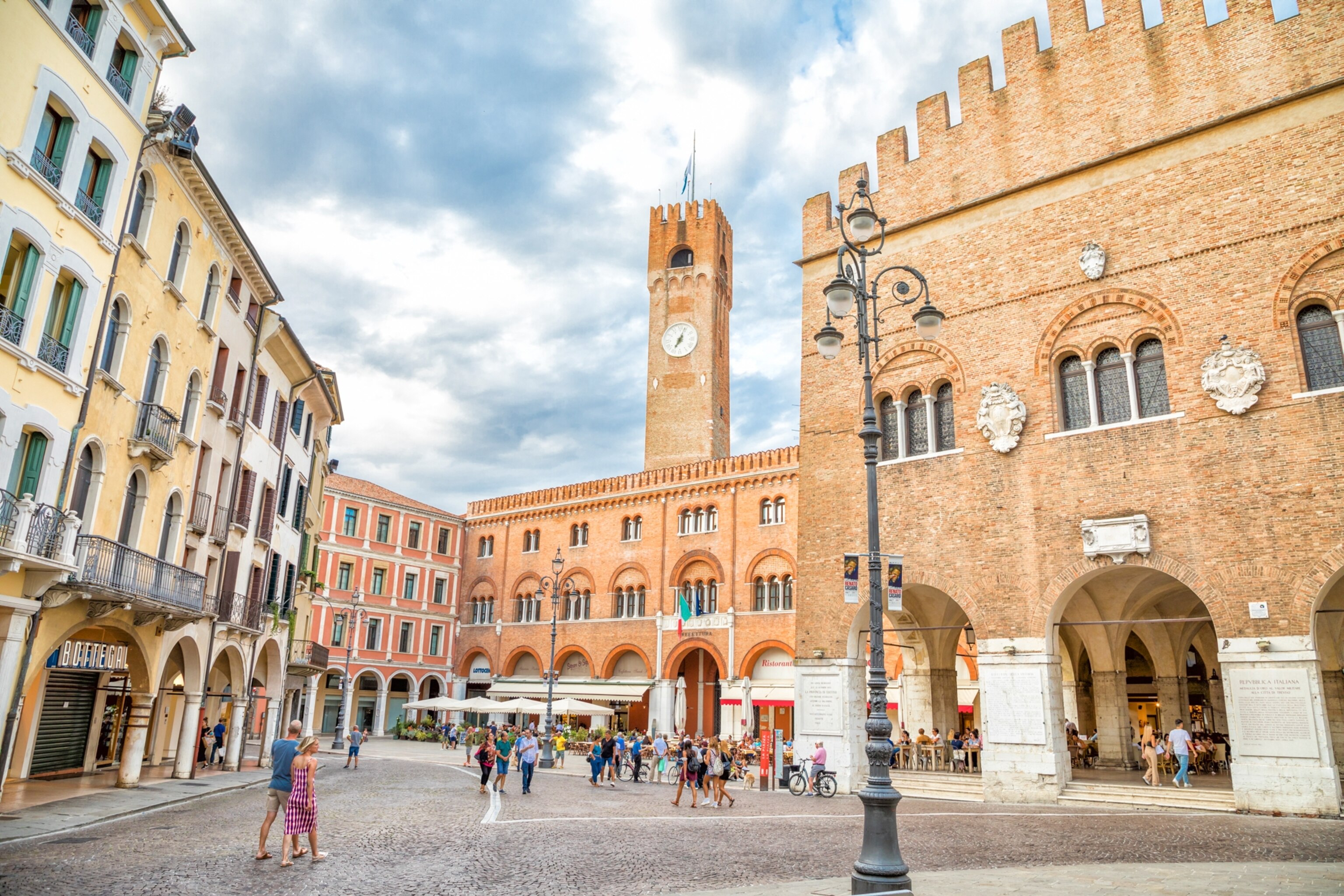 People walk across a plaza lined with stones a clock tower in the backdrop rises above the buildings that line the streets.