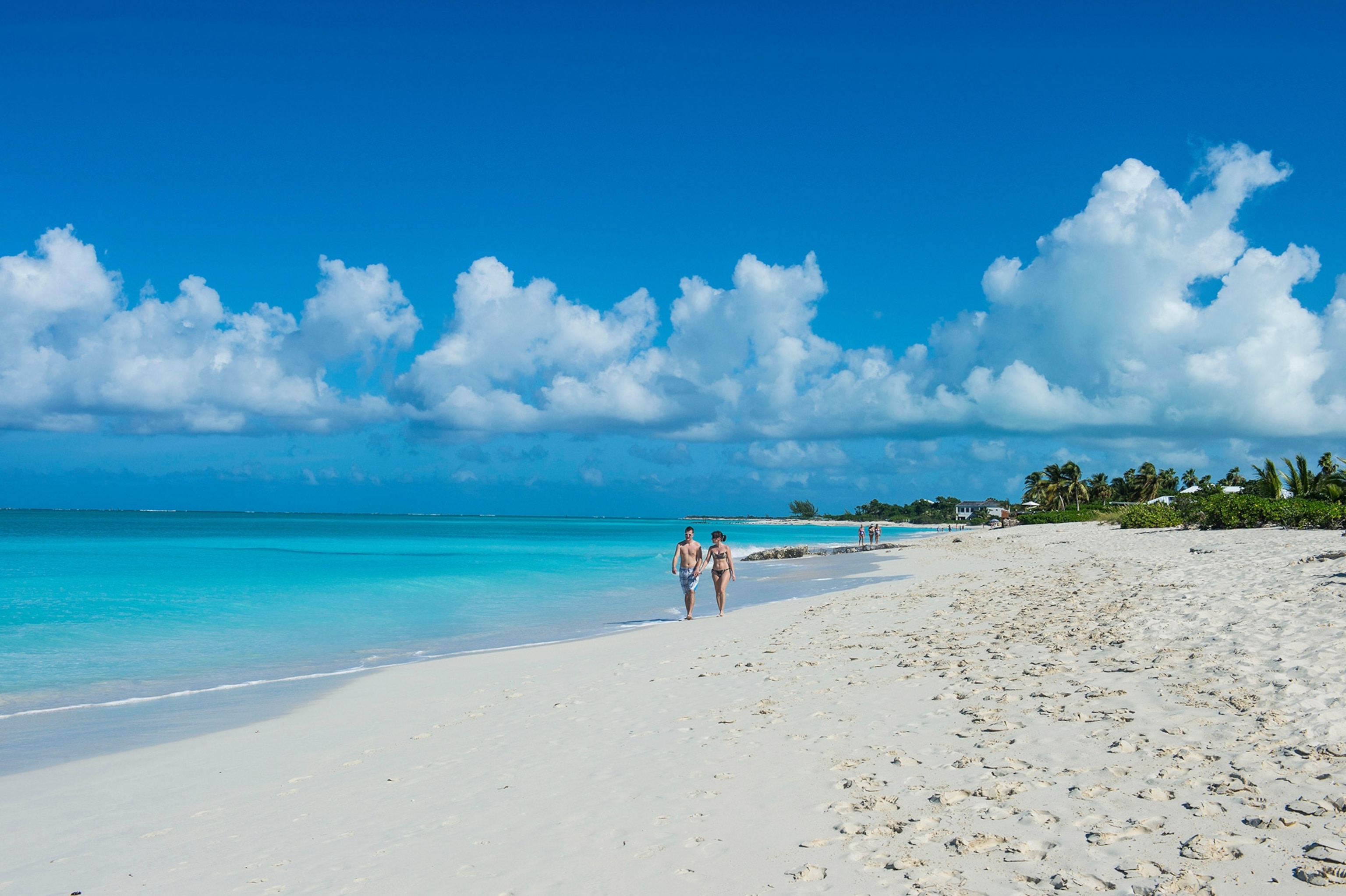 A white sand beach stretches out, clear, light blue water to the left, and green trees to the right. A couple wearing swimsuits in the distance, central frame, walks along the waterline.
