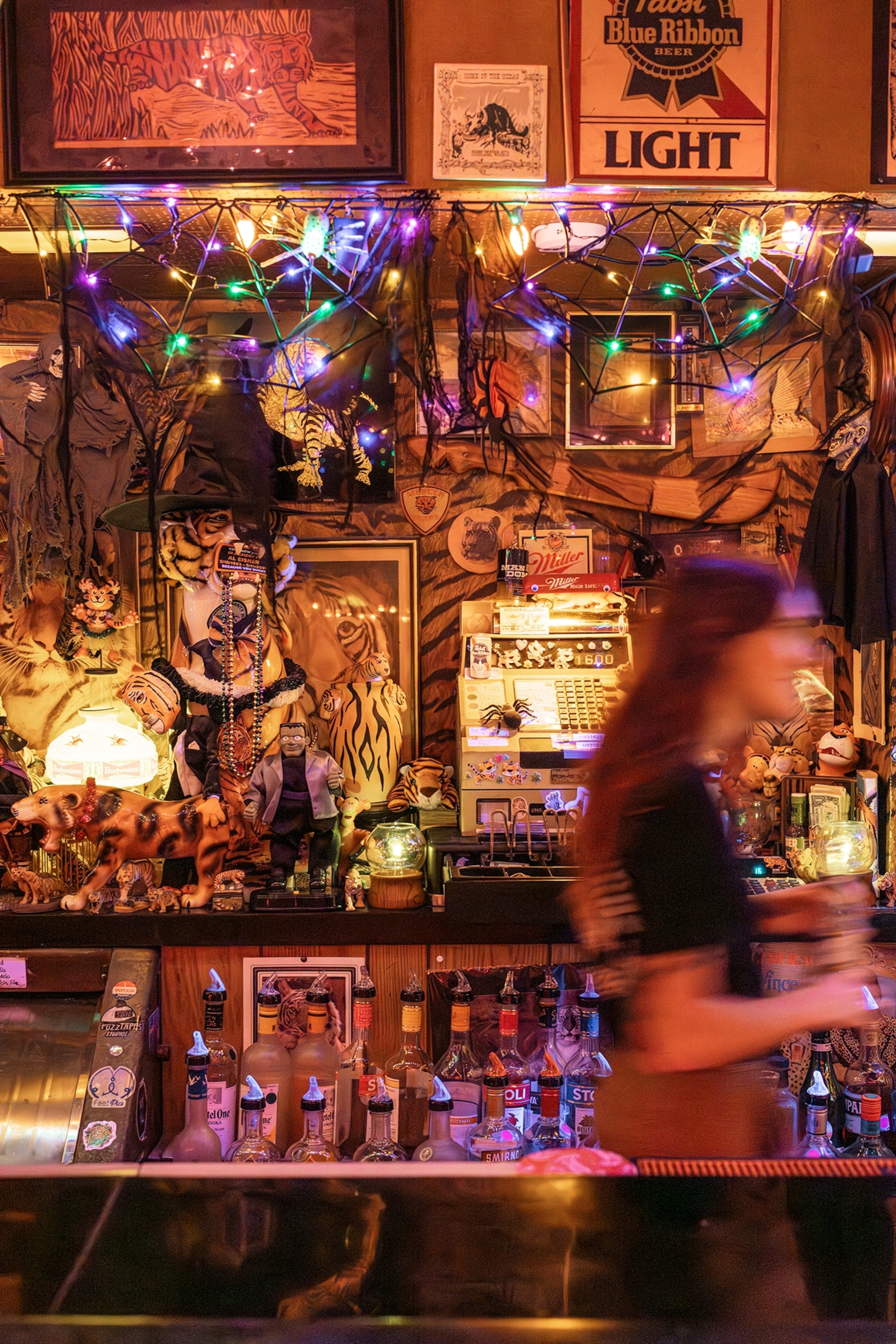 A warmly lit bar features shelves packed with vintage signs, colorful string lights, animal figurines, framed artwork, bottles, and collectibles, while a blurred person walks past the counter in the foreground.