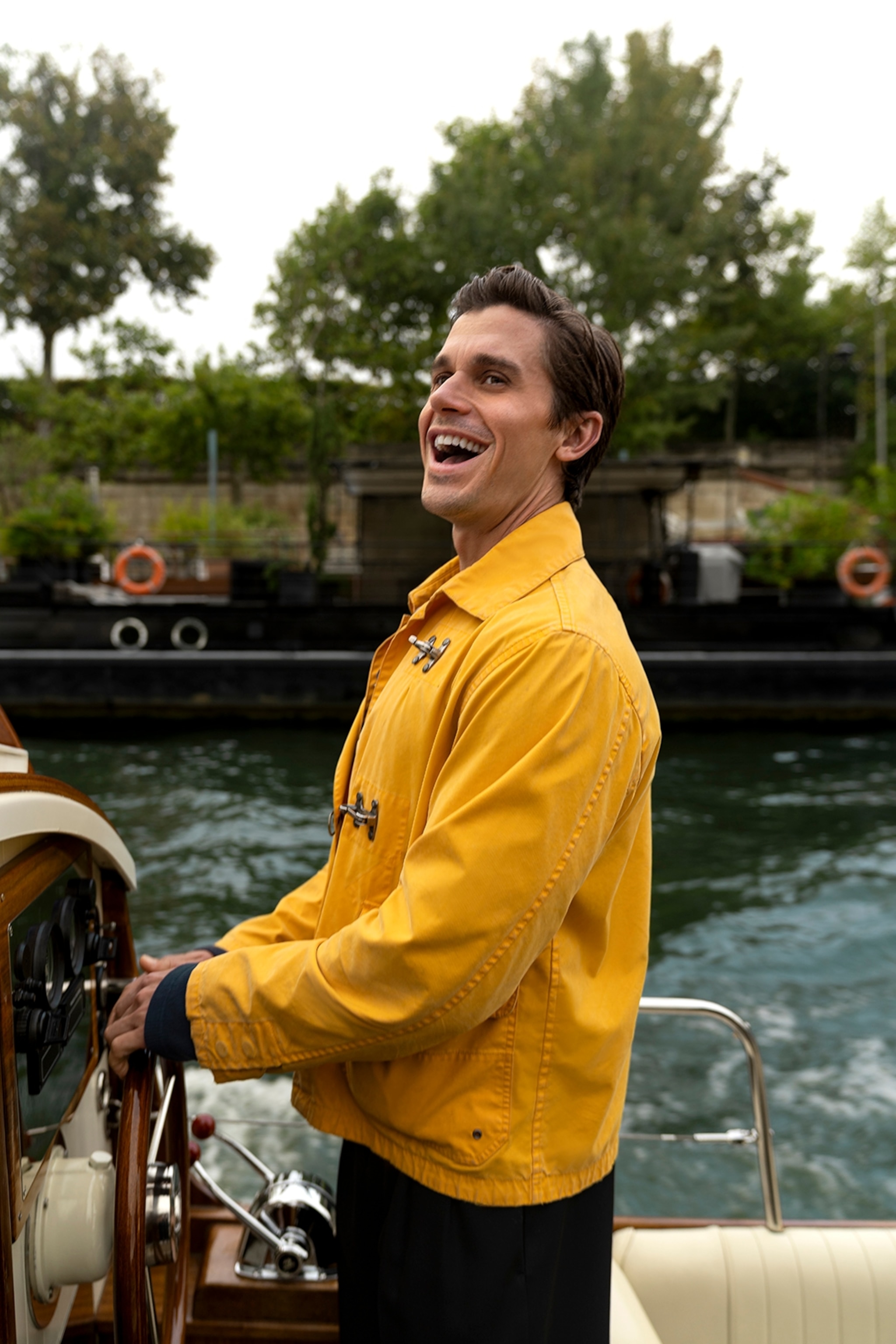 A young man in a raincoat laughing aloud while steering a canal boat.