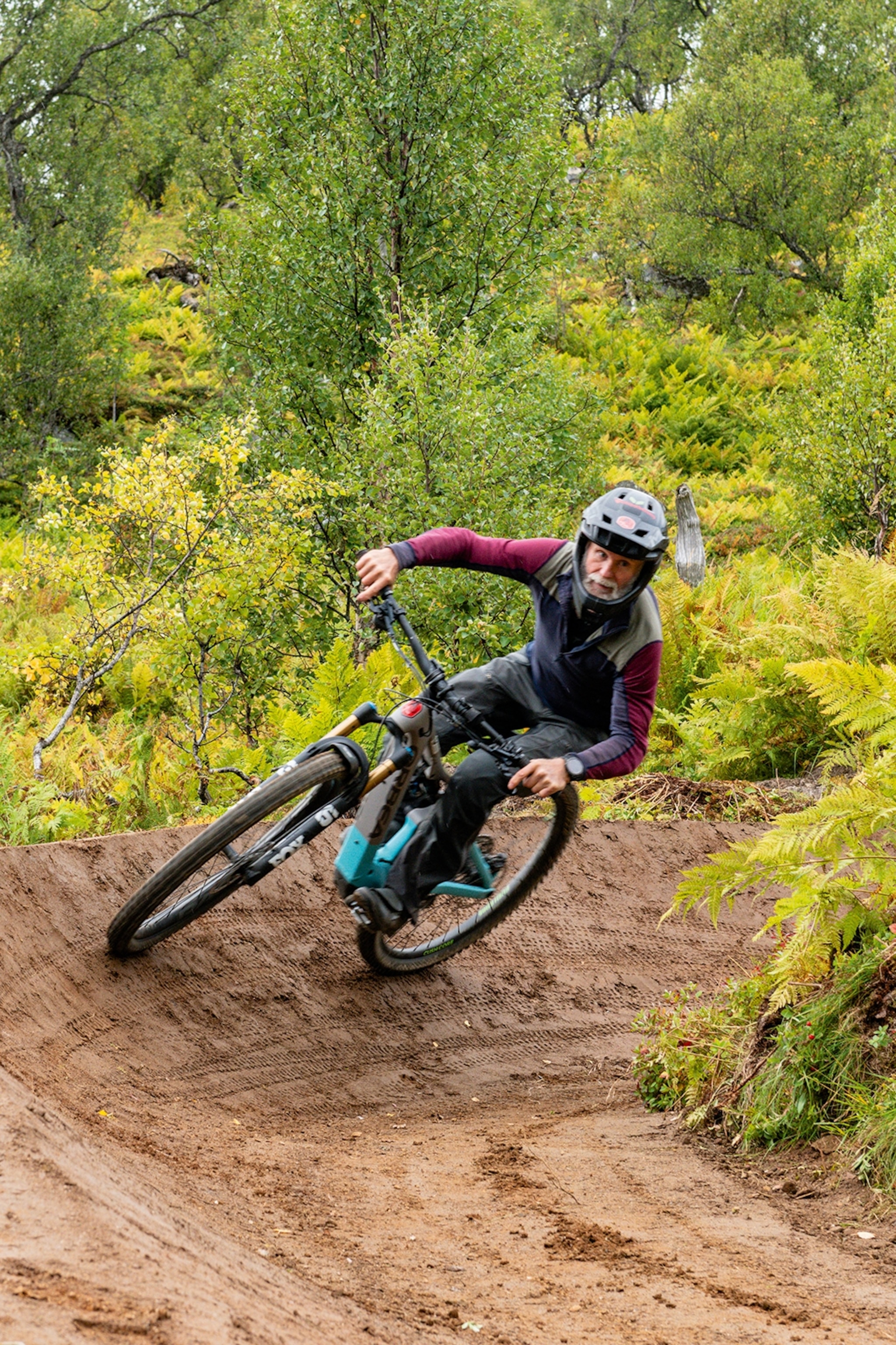 A man with white beard going around a tight corner in a natural bike park.