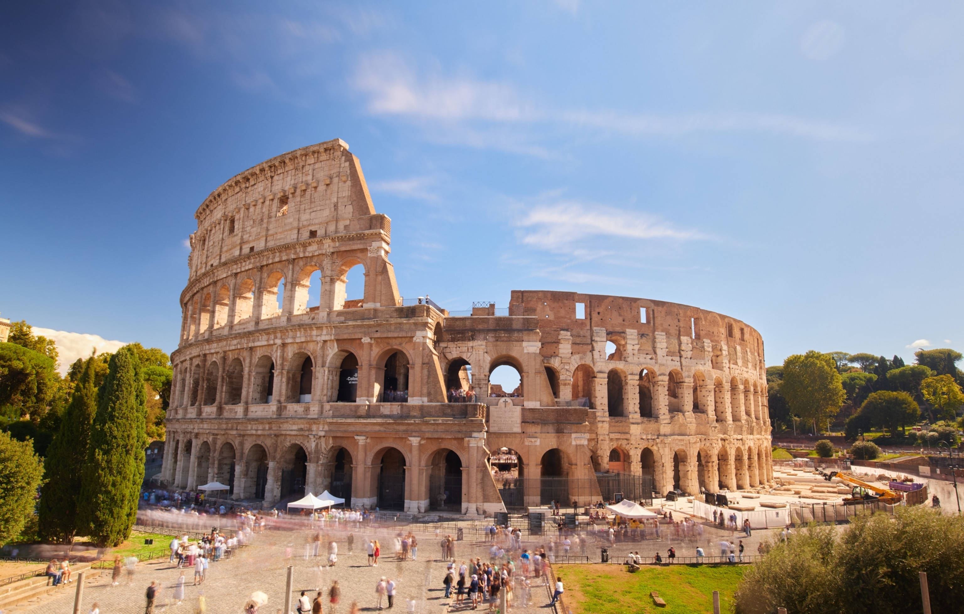 The Colosseum stands under a clear blue sky, with visitors gathered around the ancient stone amphitheater in the heart of Rome.