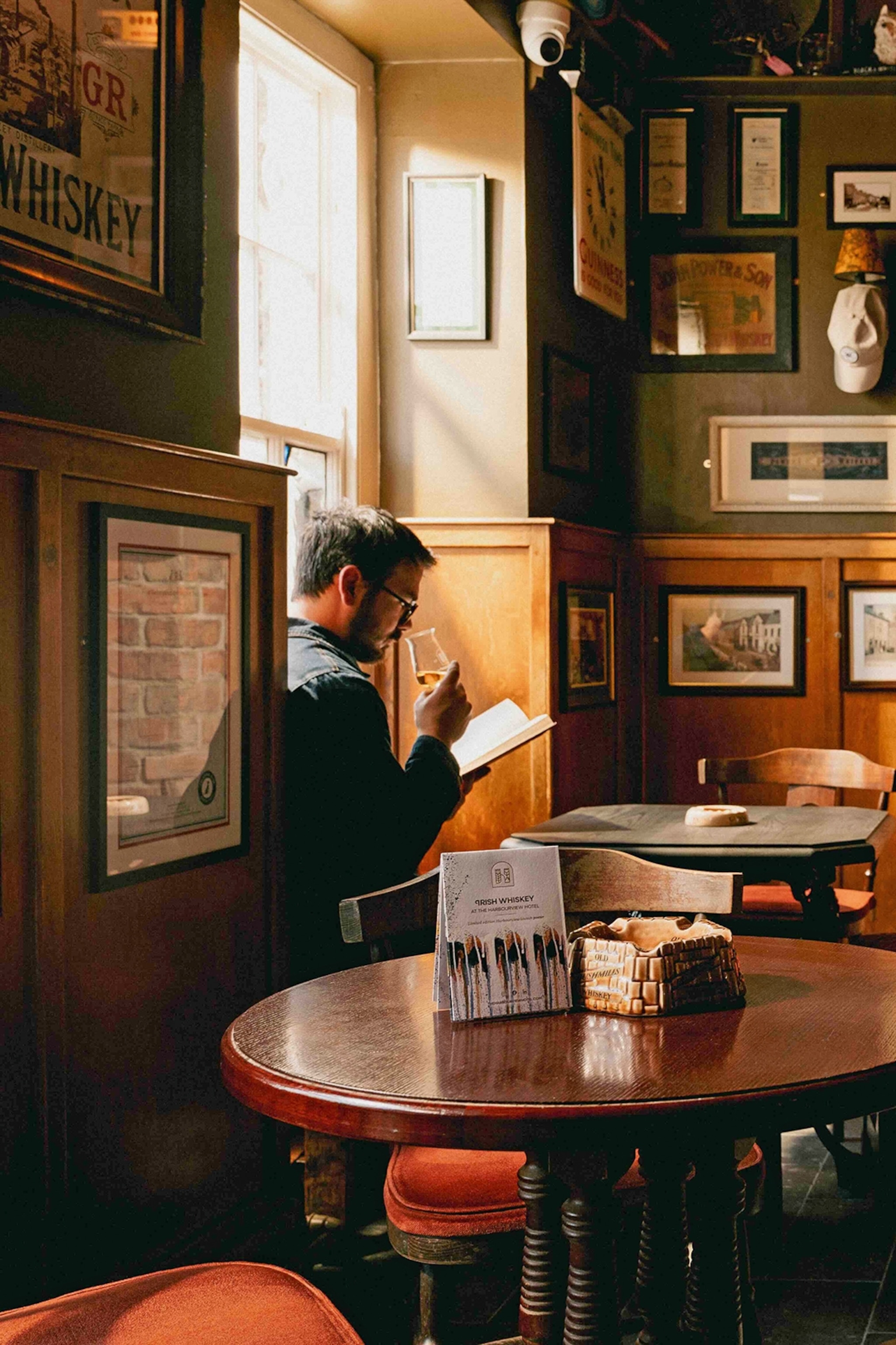 The quirky and wooden interiors of a bar with the walls donning a variety of framed pictures and a man sitting by a window enjoying a glass of whiskey.
