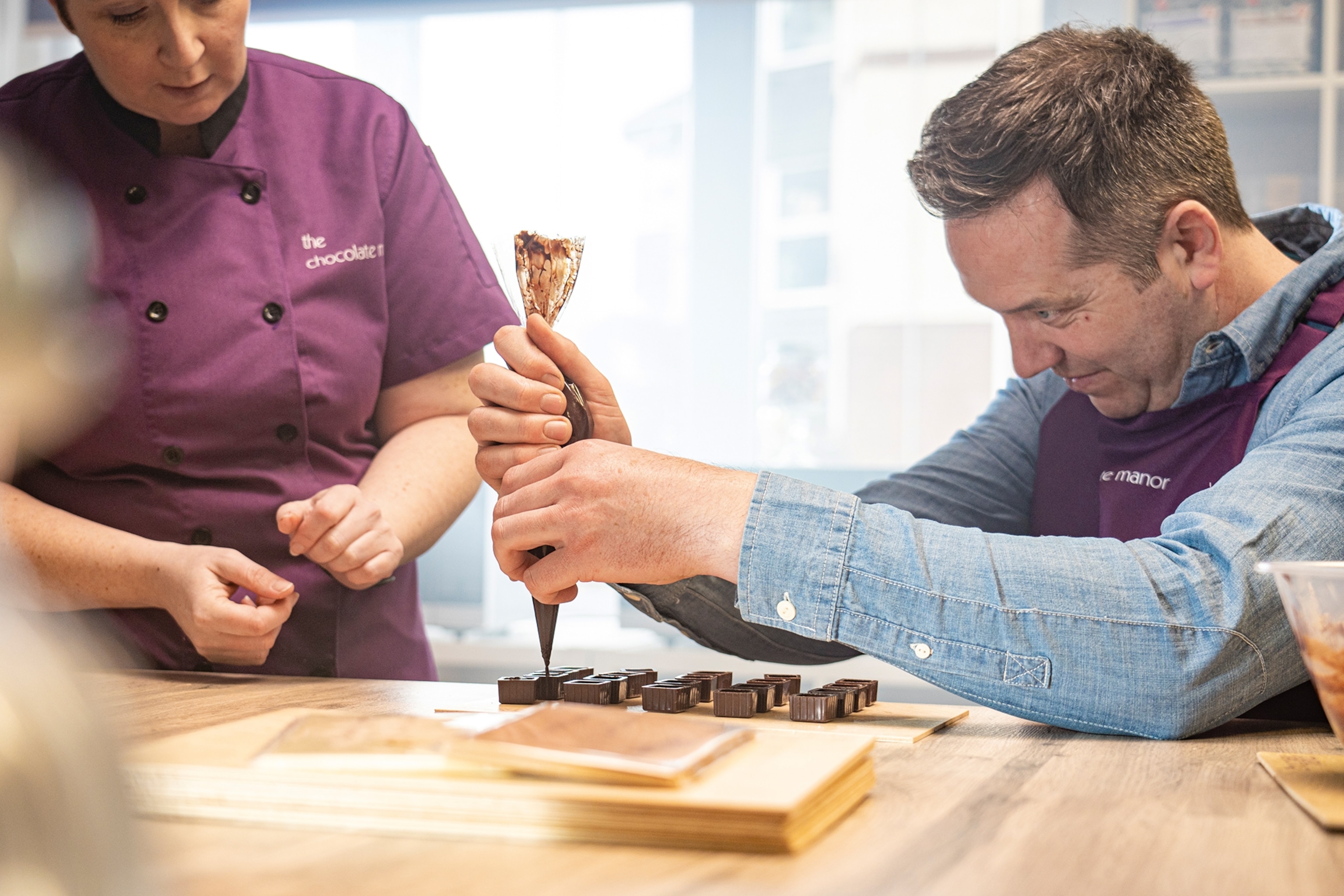 A dynamic shot taken from the height of a wooden tabling as a middle-aged man is piping chocolate onto a board.