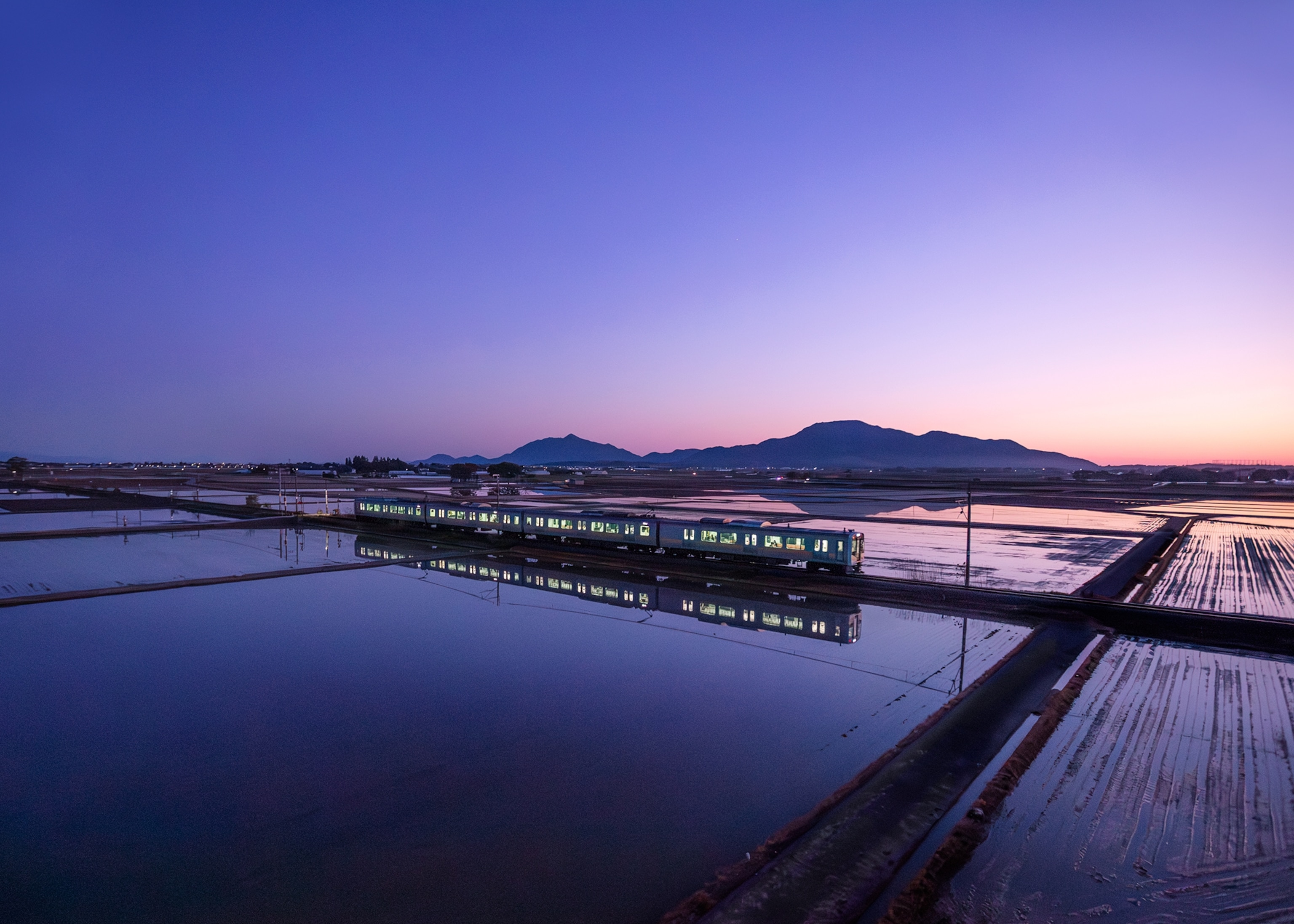A train travels through flooded rice fields at dusk, with its lights reflected in the still water and mountains silhouetted against a purple and pink evening sky.