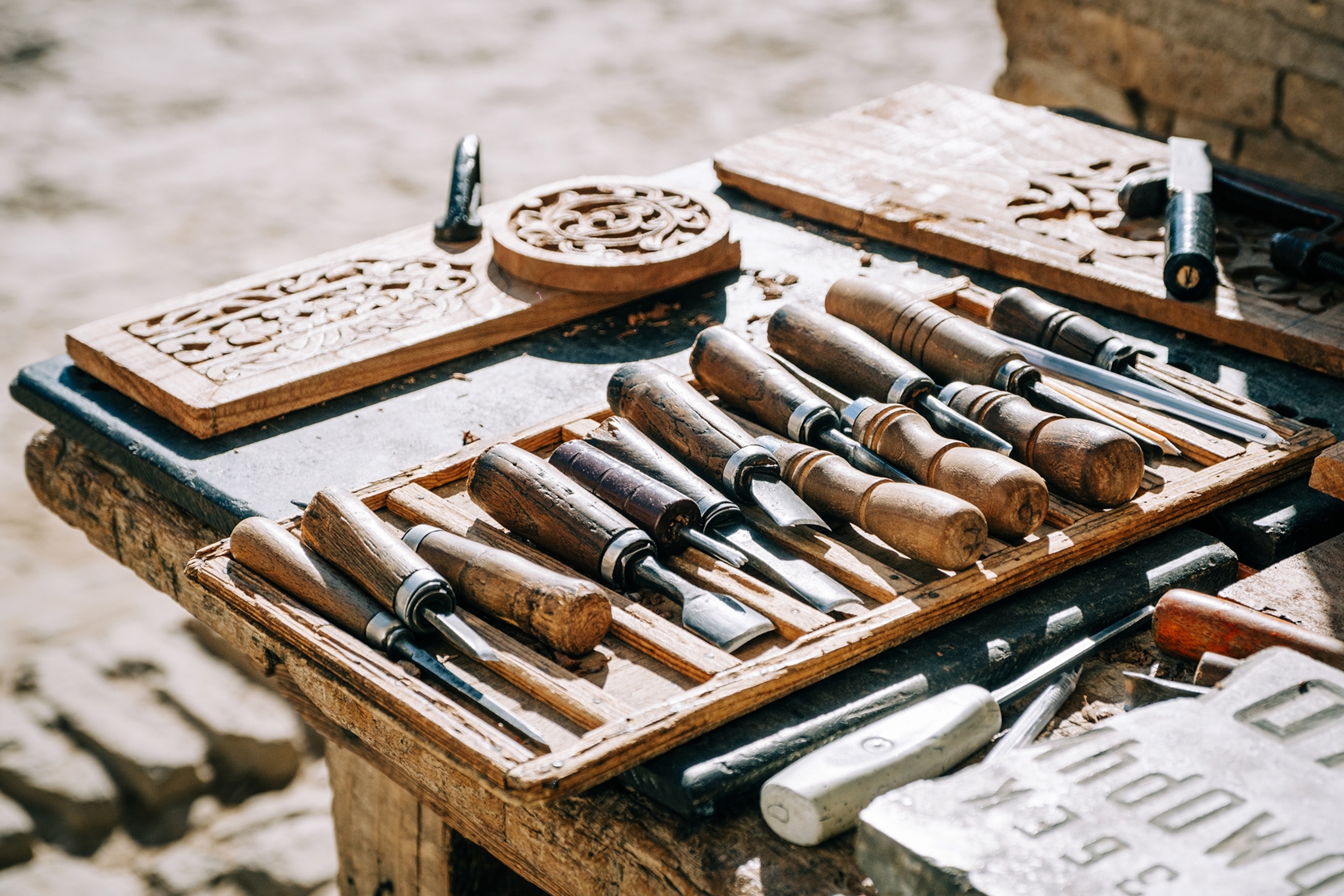 A close-up shot of an old wooden table, where a selection of carving tools is neatly laid out and illuminated by the sun.