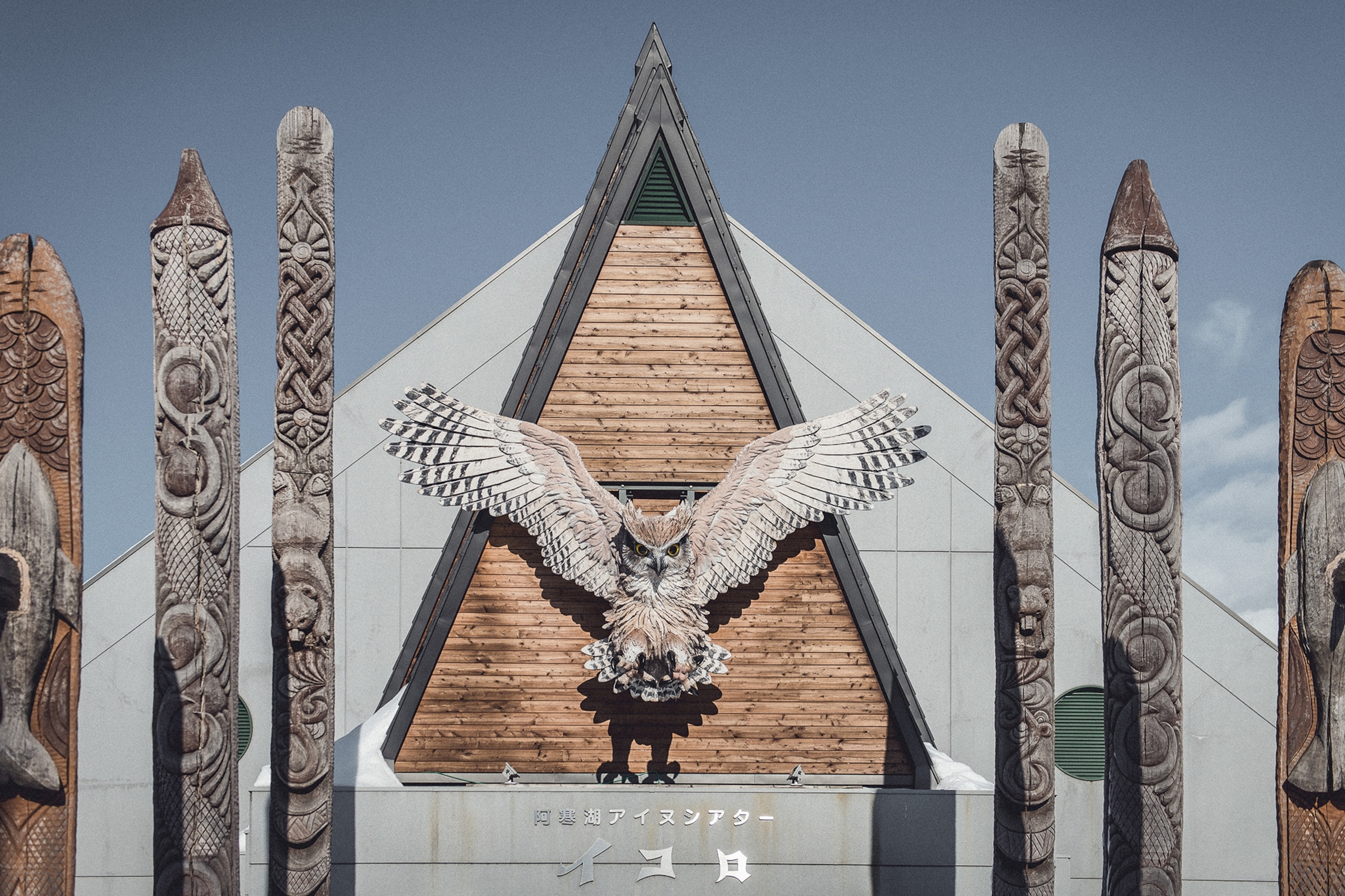 The wooden facade of a tent-like building with carved totem poles on either side of the entrance and a carved, wooden owl towering above.