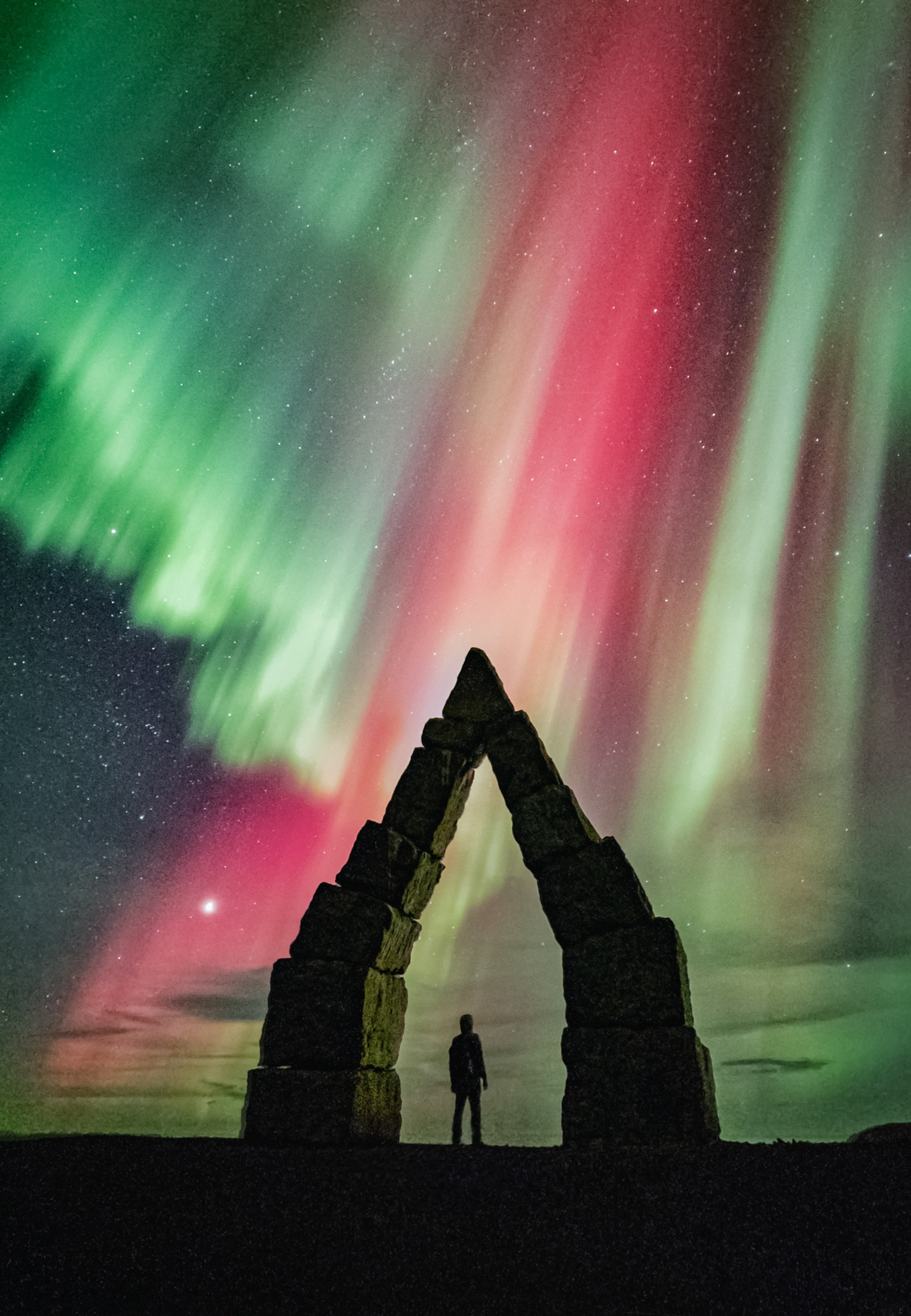 A silhouetted figure stands beneath a stone archway, gazing at the vibrant green and red aurora borealis illuminating the starry night sky