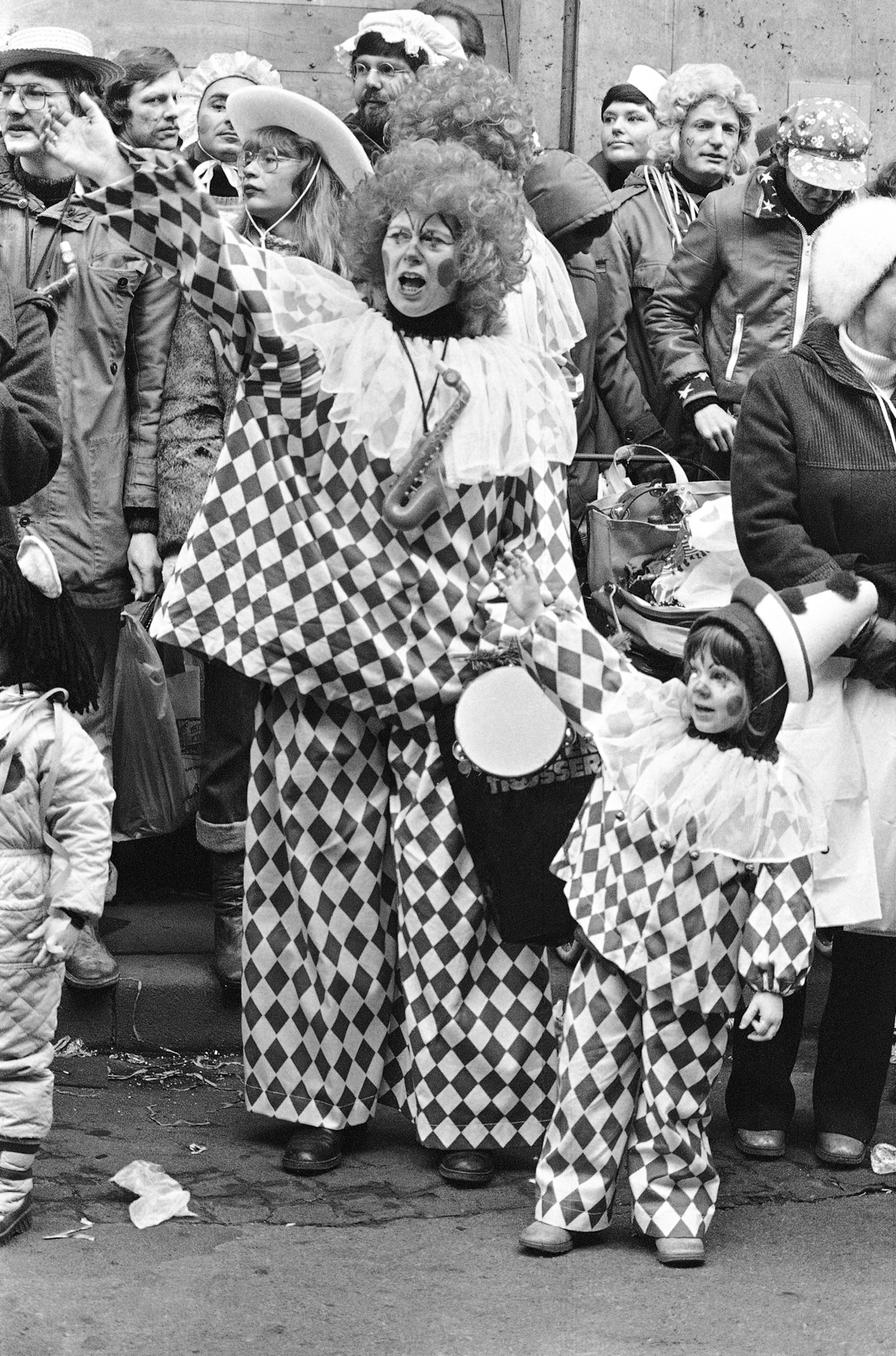 A black and white photograph of a mother and child in clown checkered costumes amongst a group of people.