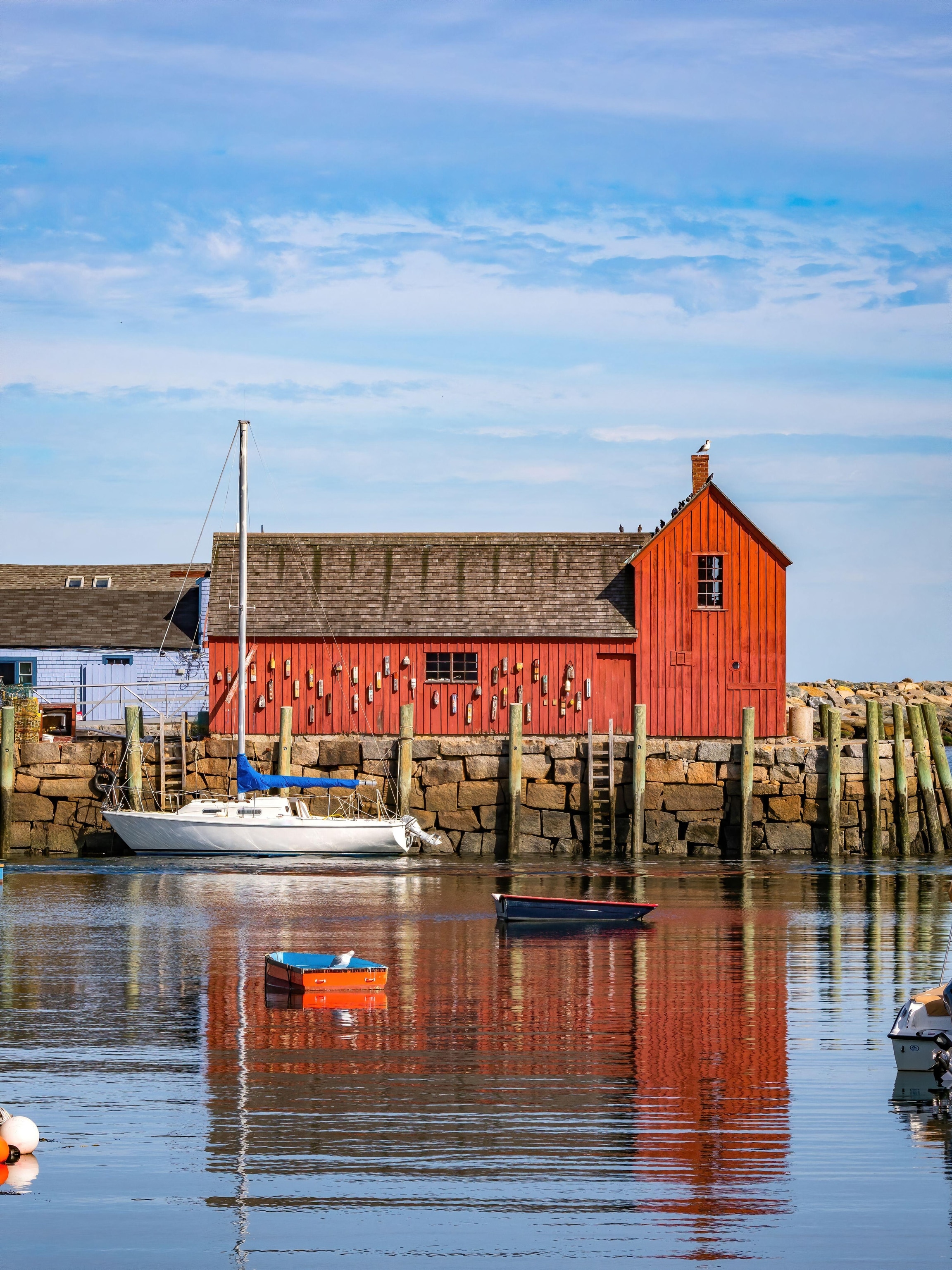A red fishing shack sits on an island in on the ocean, it's reflection bright in the water with boats passing by.