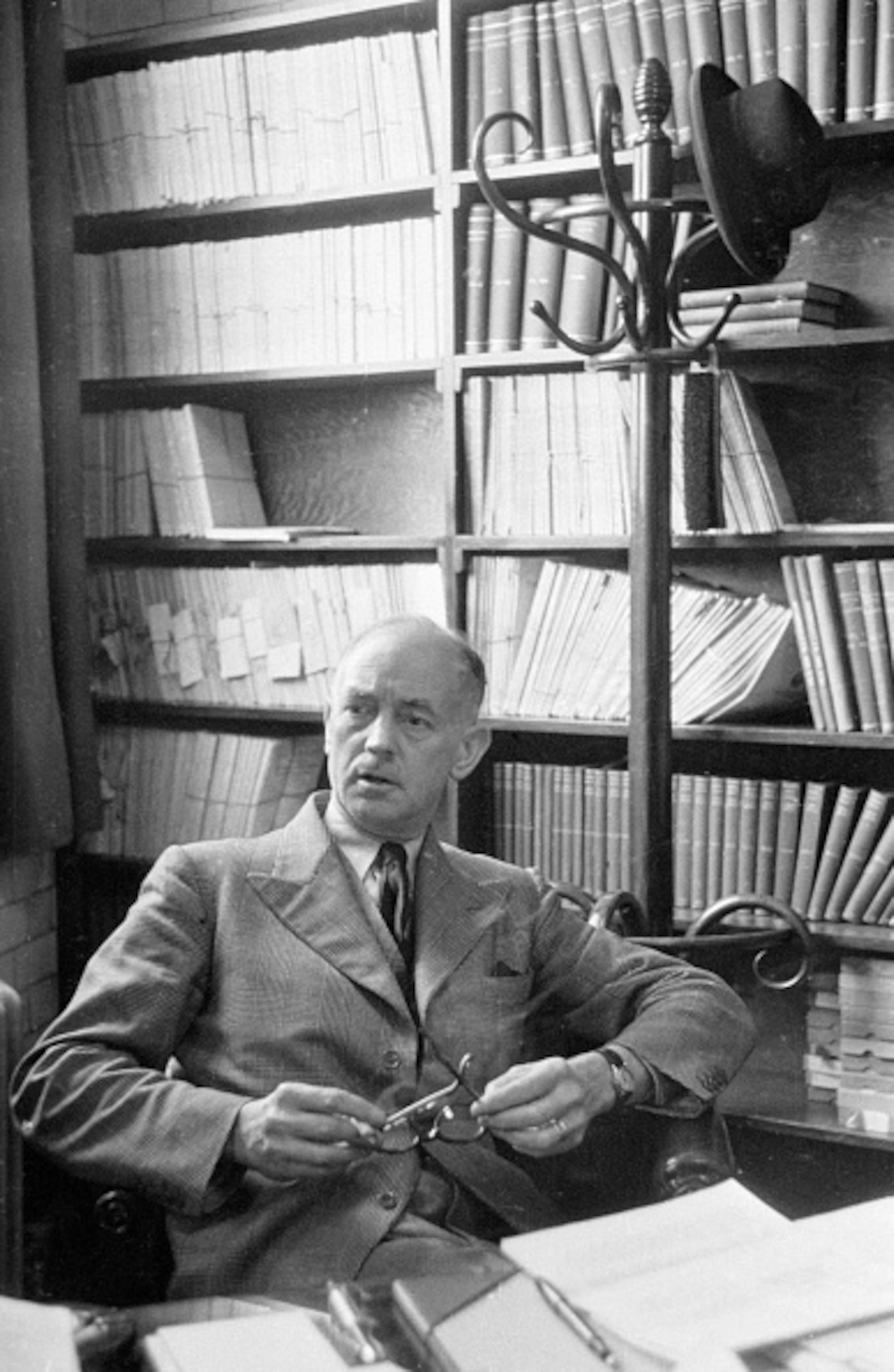 Black and white portrait of a man in a suit and tie, sitting behind a paper-strewn desk, holding a pair of eyeglasses. Behind him a large bookshelf and a tall hat rack with a hat hanging atop.
