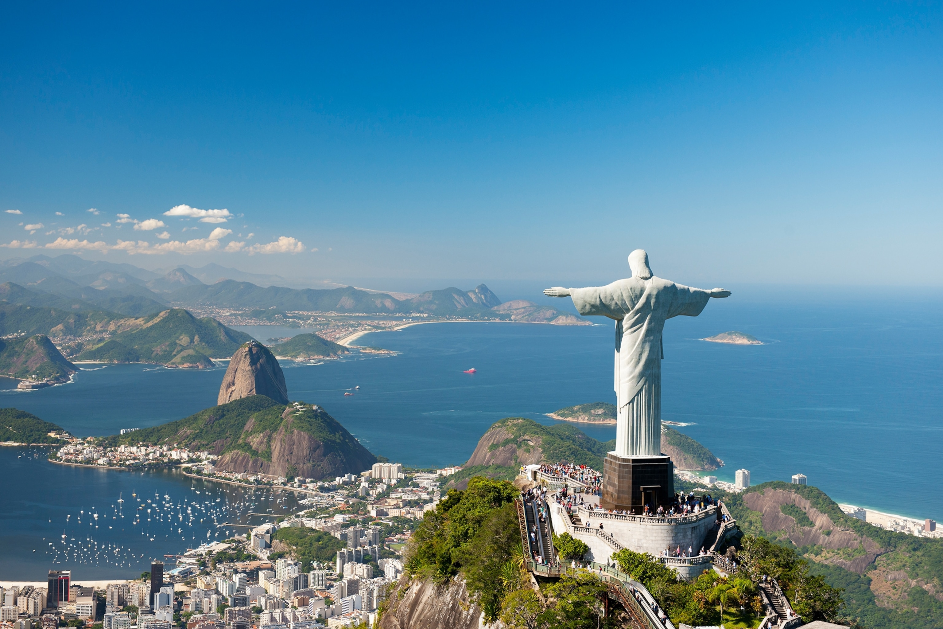A statue of Christ with open arms at the top of a hill, overlooking a seaside city in Brazil.