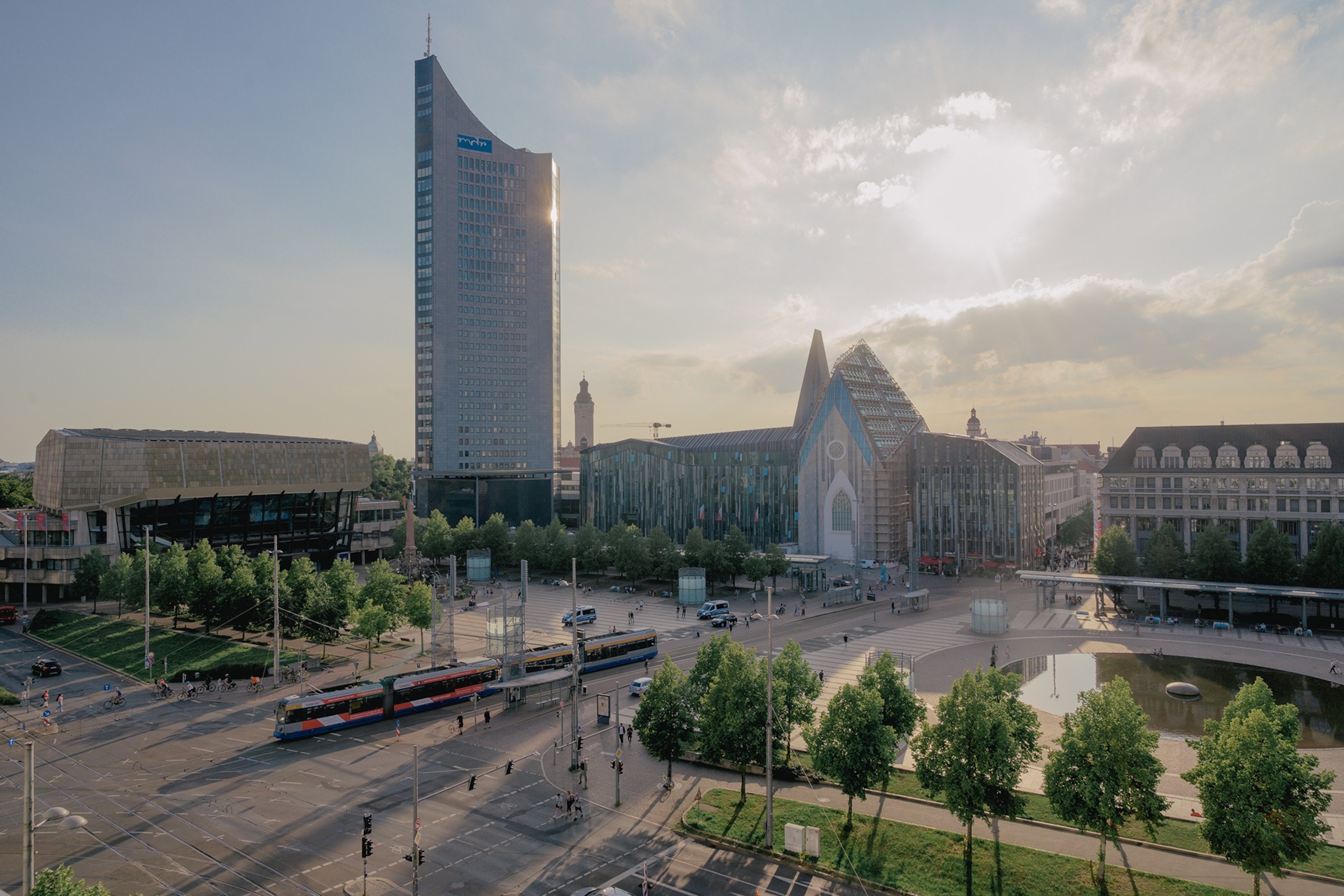 A modern city square features a tall glass skyscraper, contemporary buildings, tram tracks, and tree-lined streets, with sunlight filtering through clouds above the urban landscape.
