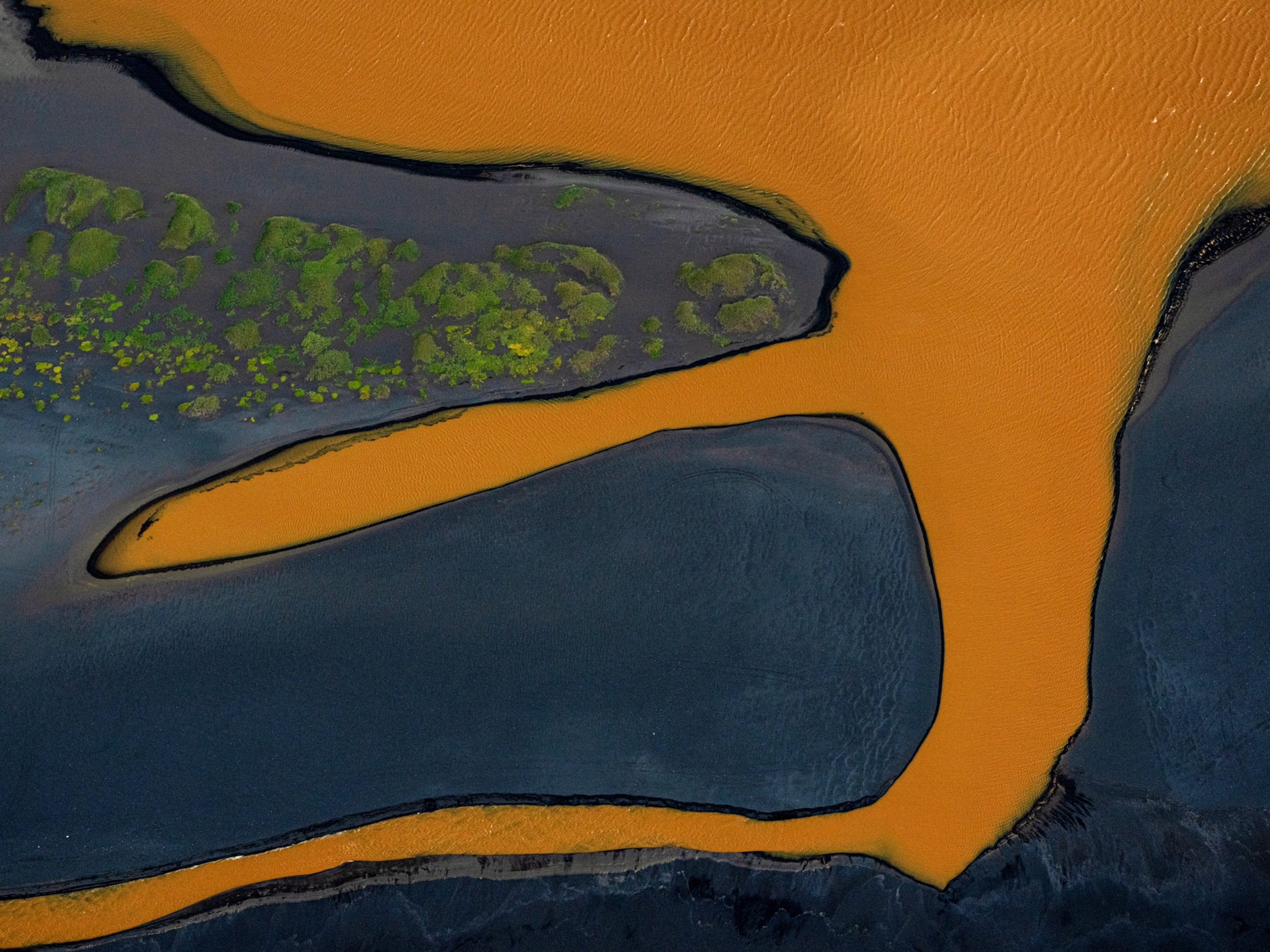 Aerial view of an abstract landscape featuring bright orange water curving against dark patches of land dotted with vibrant green vegetation