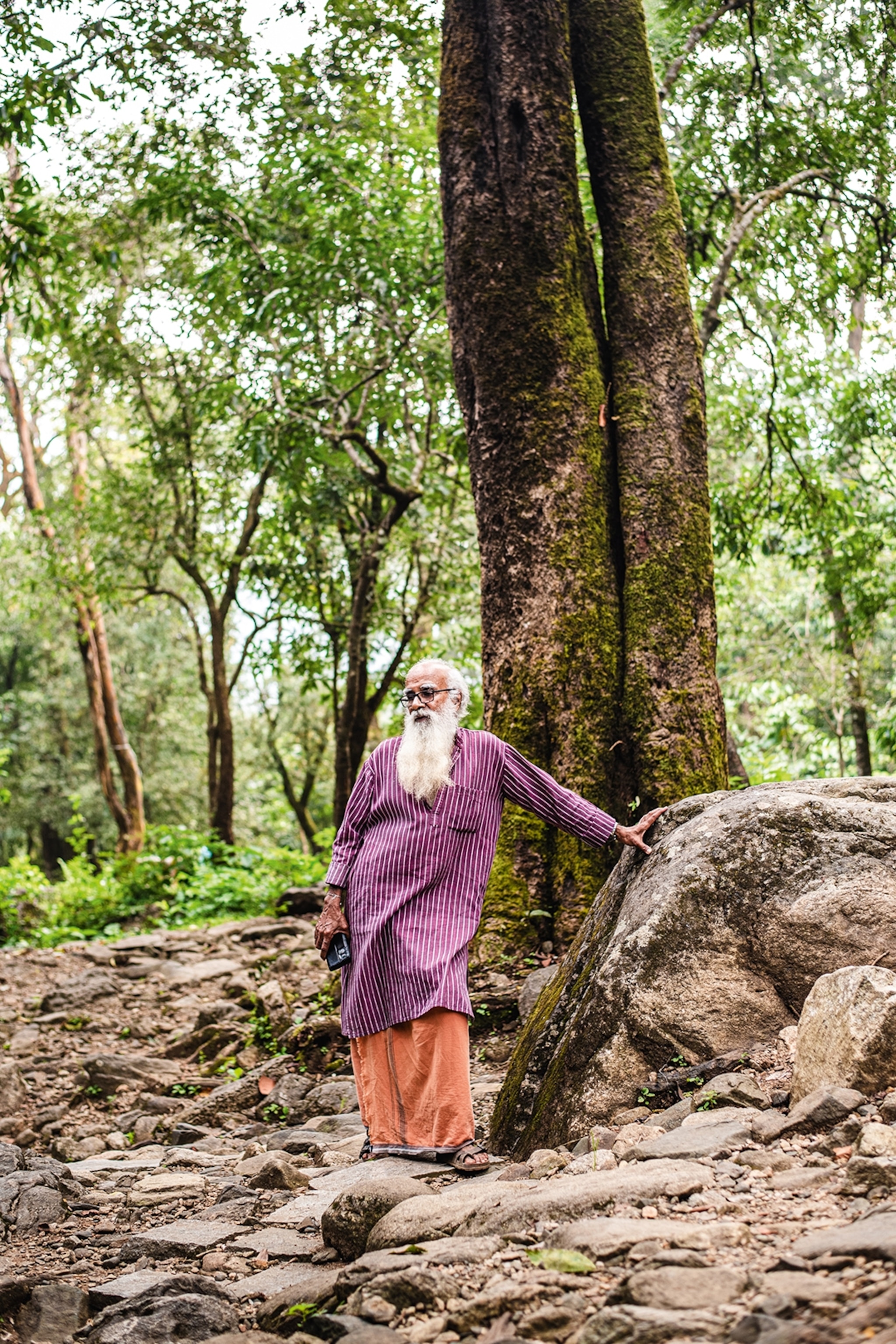 An elderly man with a long, wispy beard seemingly lost in thought as he's leaning with his hand against a rock in a forest.